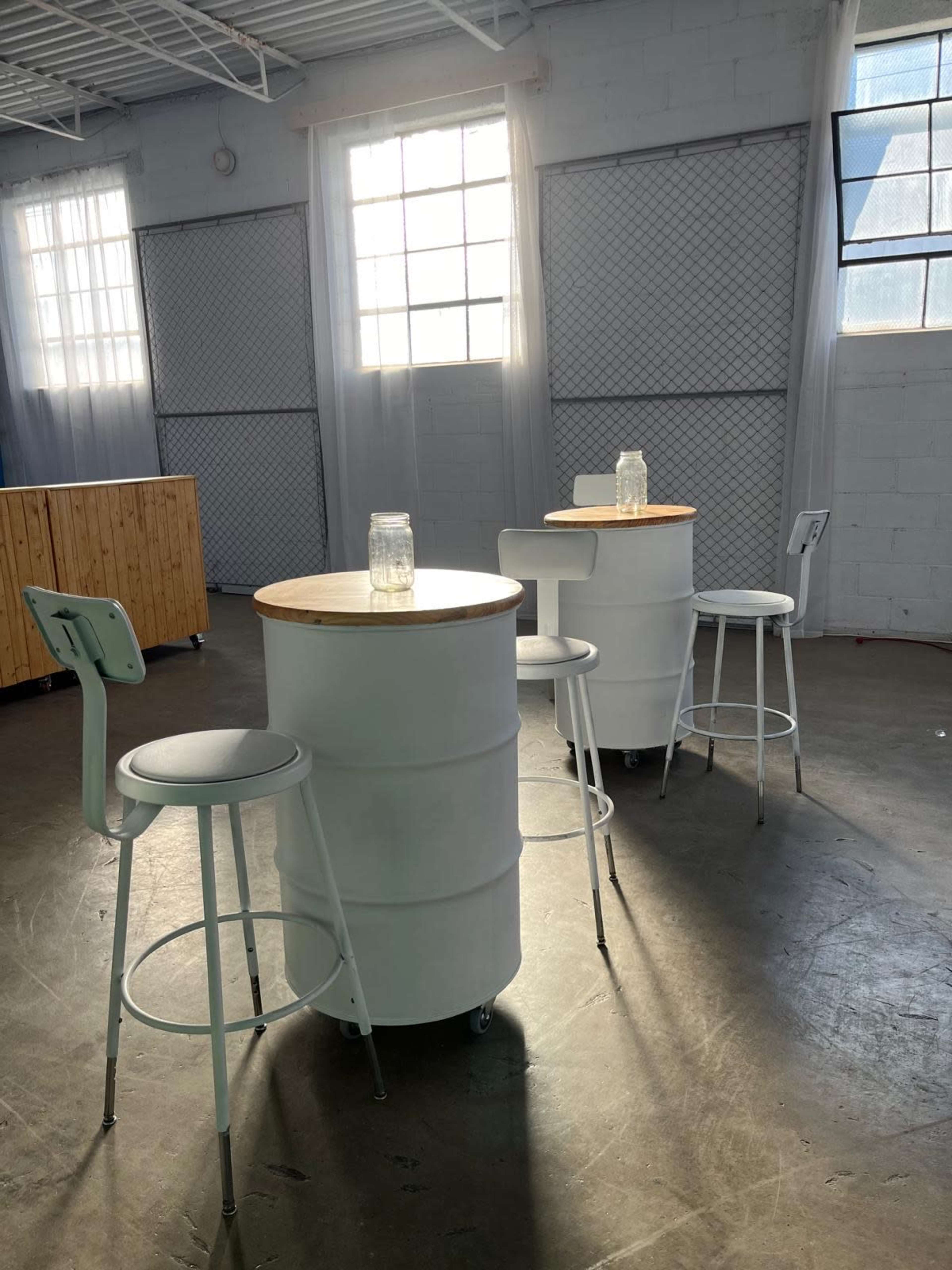The image shows a modern indoor space featuring round tables made from white barrels with wooden tops and light-colored stools.