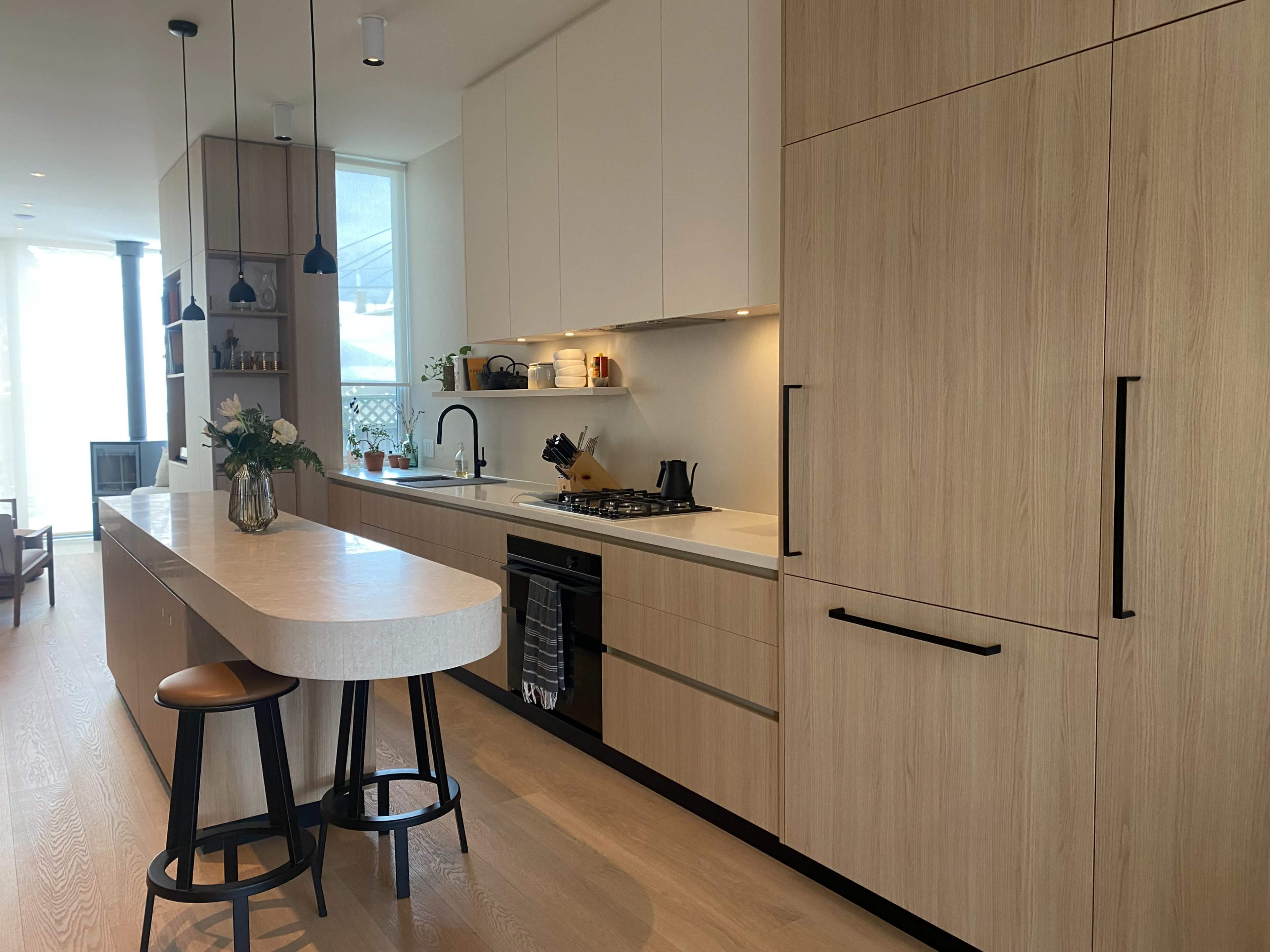 The image shows a modern kitchen with wooden cabinetry, a large island with bar stools, and a stove integrated into the counter.