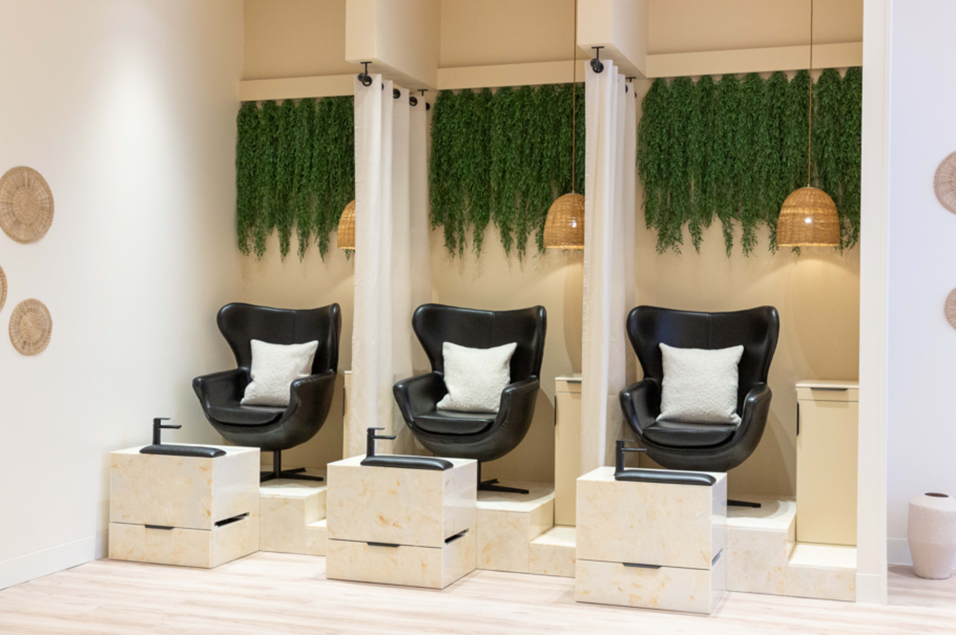 The image shows a modern salon setup with three black chairs facing a mirrored wall, surrounded by greenery and pendant lighting.