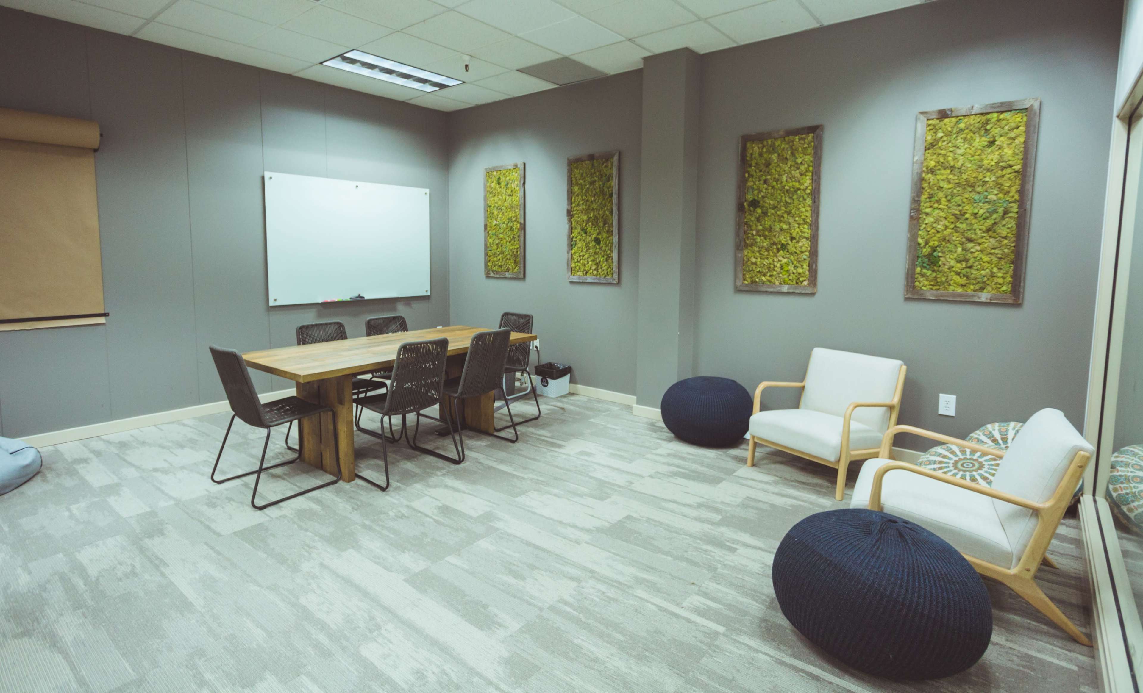 A conference room features a wooden table surrounded by chairs, with a whiteboard on one wall and moss-covered artwork on the adjacent walls.