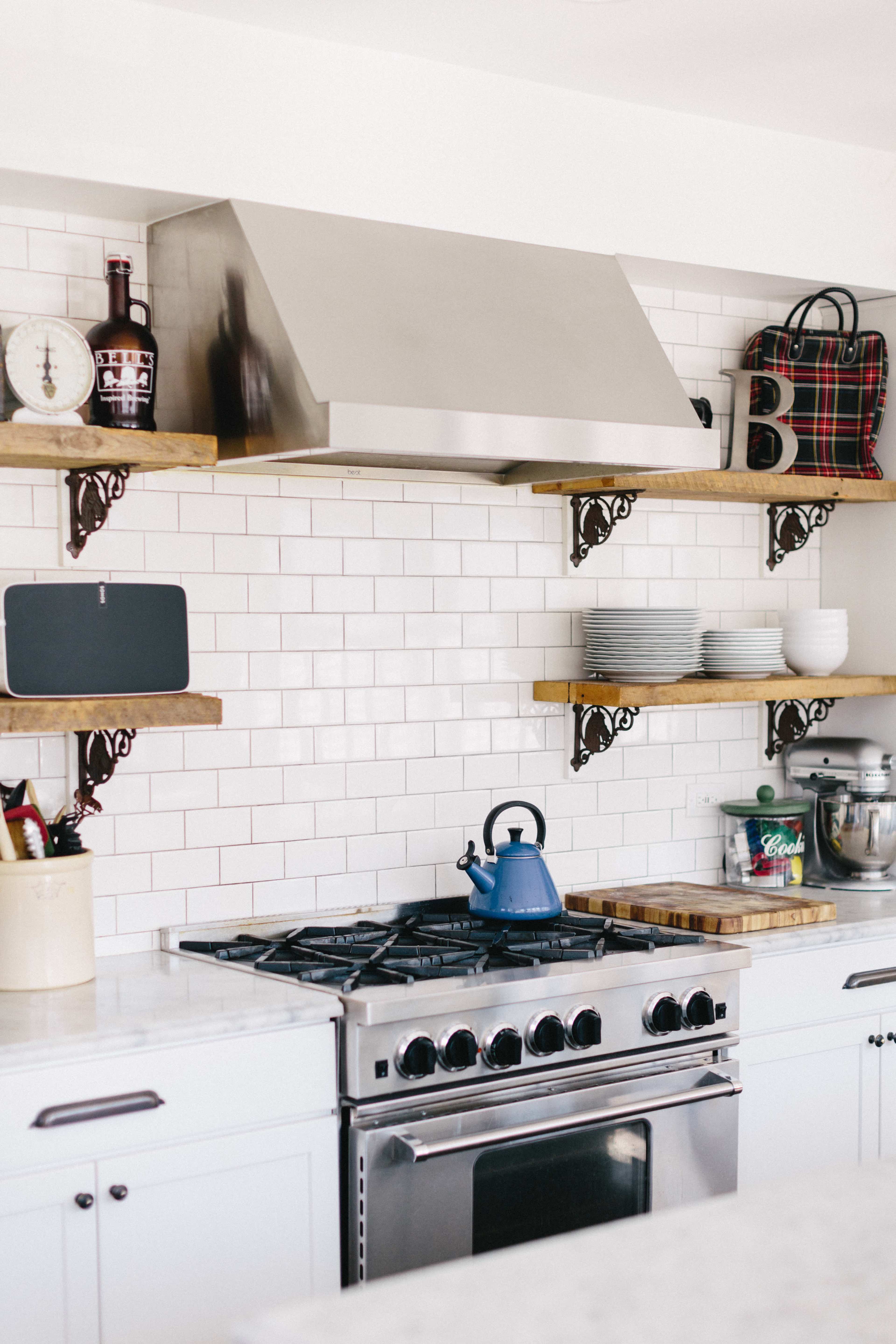 A modern kitchen features a stainless steel range hood above a gas stove, flanked by wooden shelves holding plates and kitchen accessories.