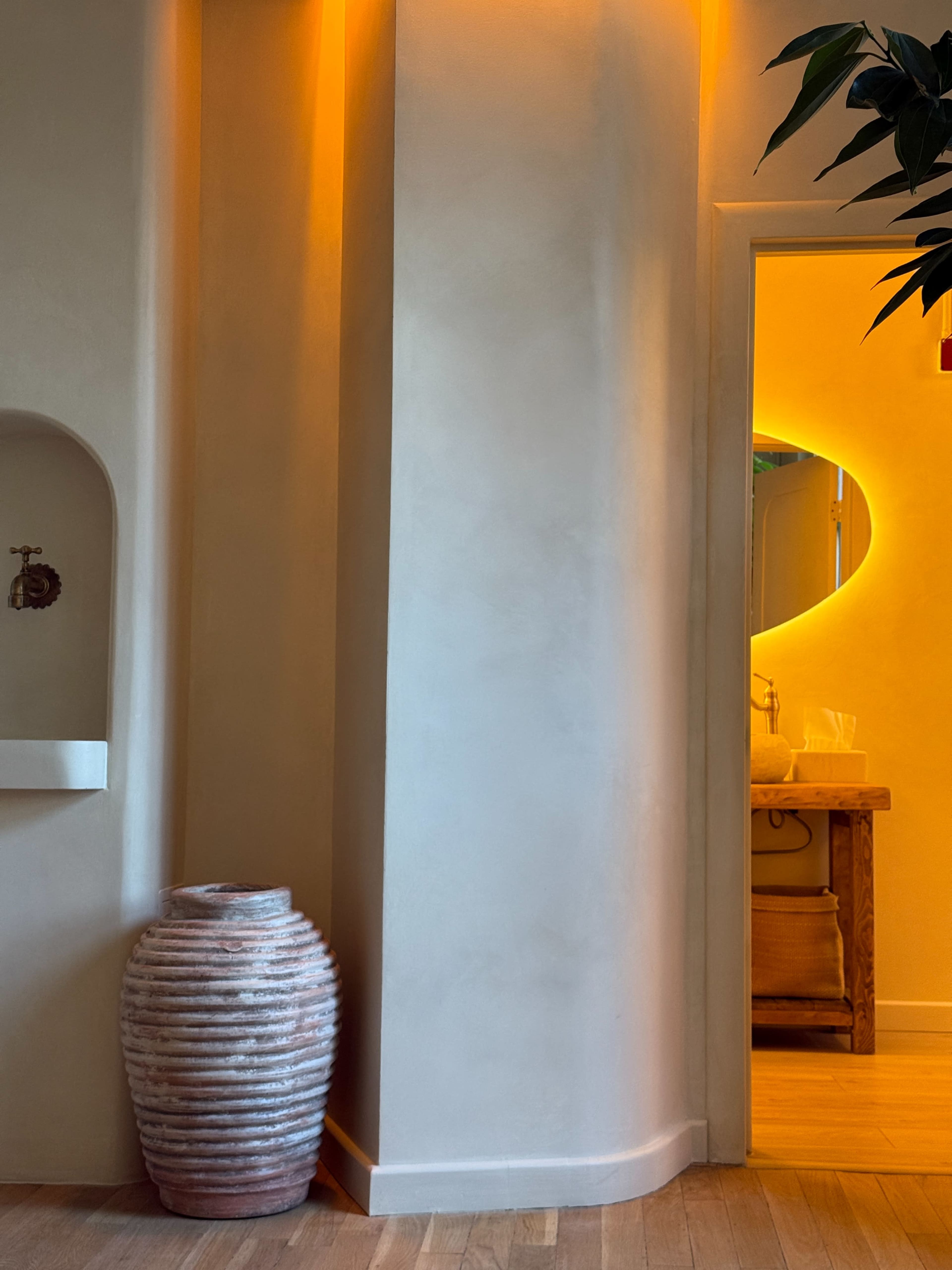 A textured vase is placed against a light-colored wall next to a softly lit doorway that leads to another room.