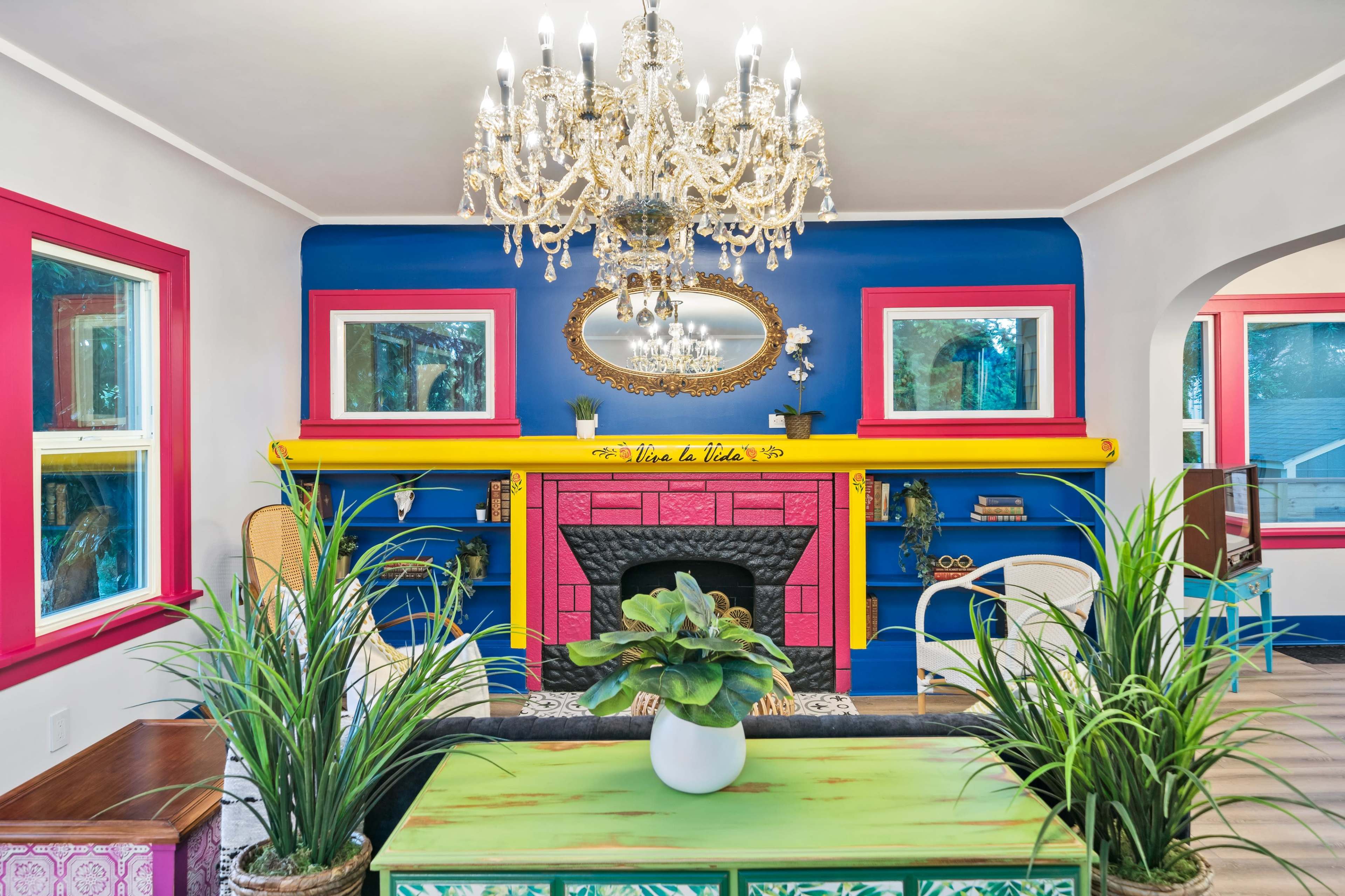 A living room featuring a colorful fireplace with a chandelier overhead, surrounded by potted plants and decorative furniture.