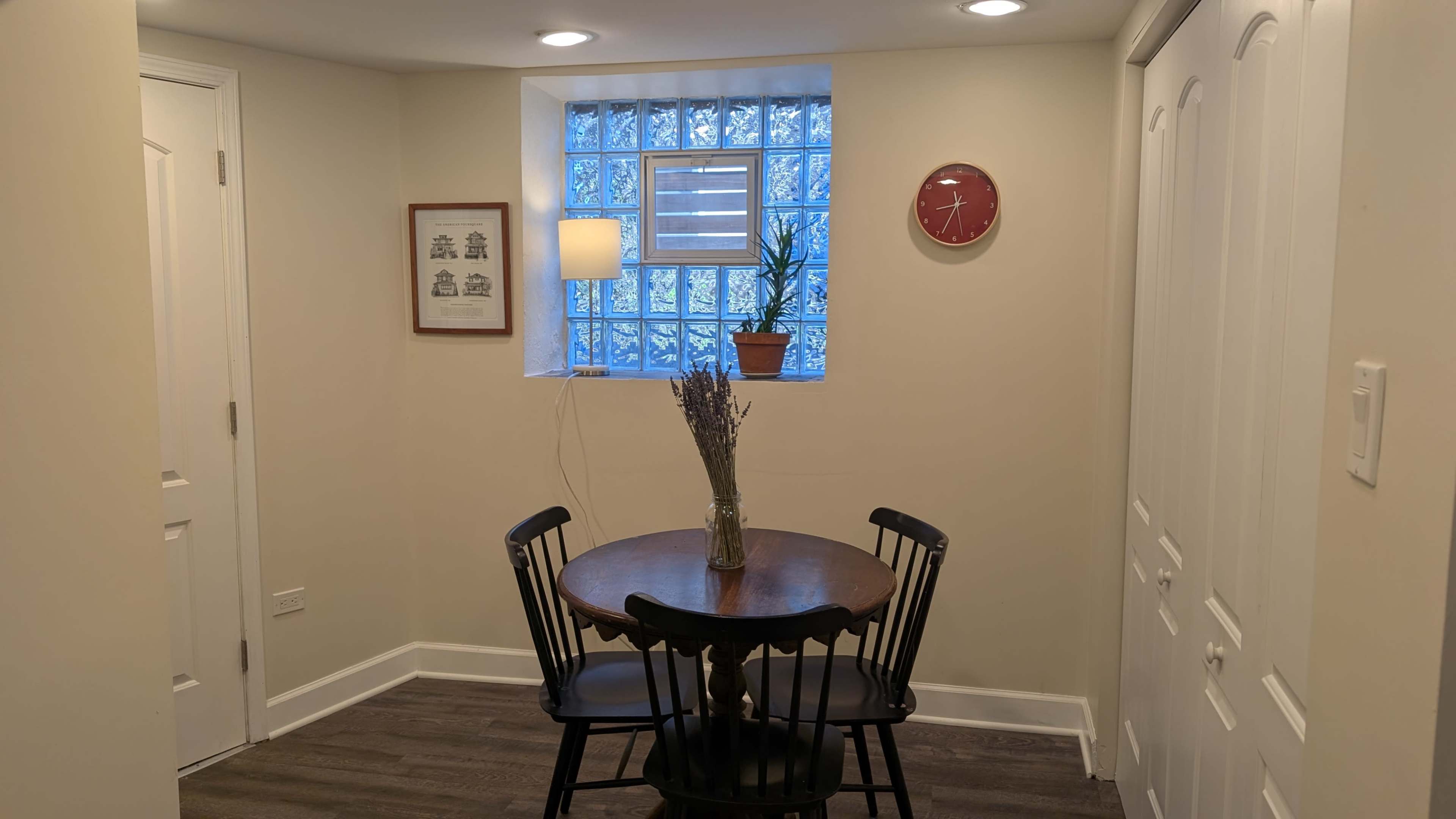 The image shows a small dining area with a round table and two black chairs, a wall clock, a plant on a windowsill, and framed artwork.