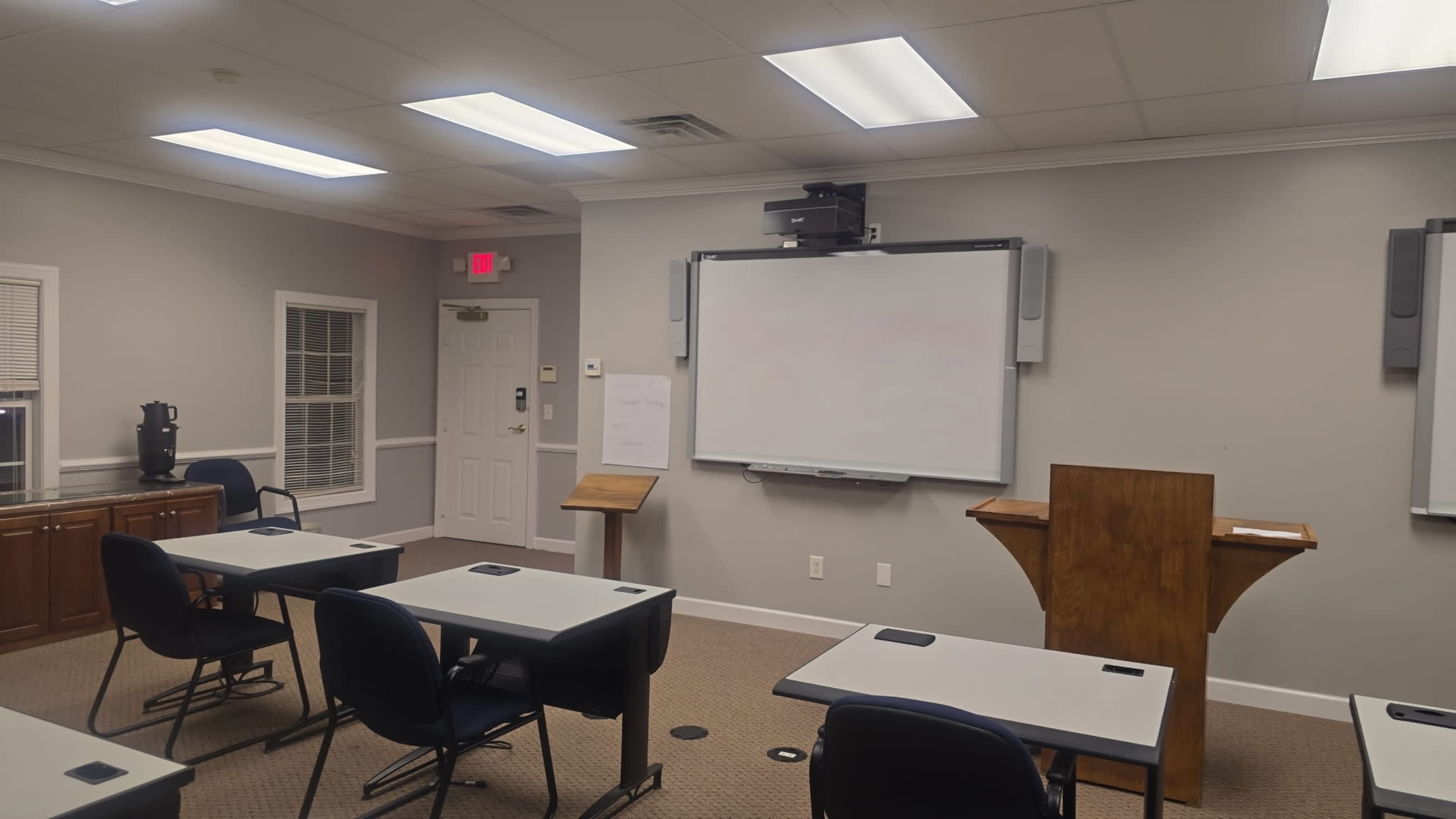 The image shows a classroom setup with empty desks facing a large whiteboard and a podium, illuminated by overhead lights.