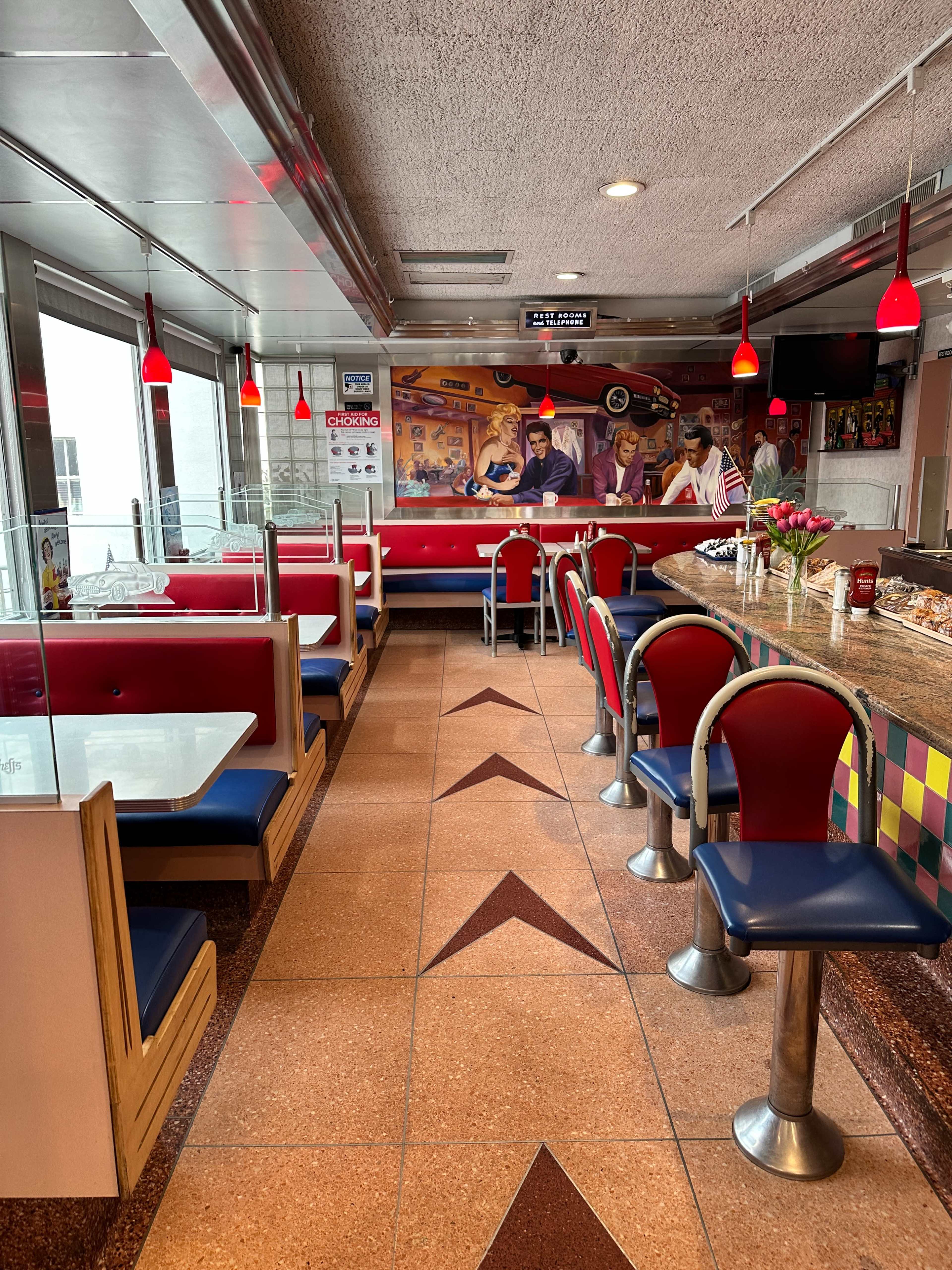 A diner interior with red and blue seating booths, a curved counter with bar stools, and a mural depicting a food theme on the wall.