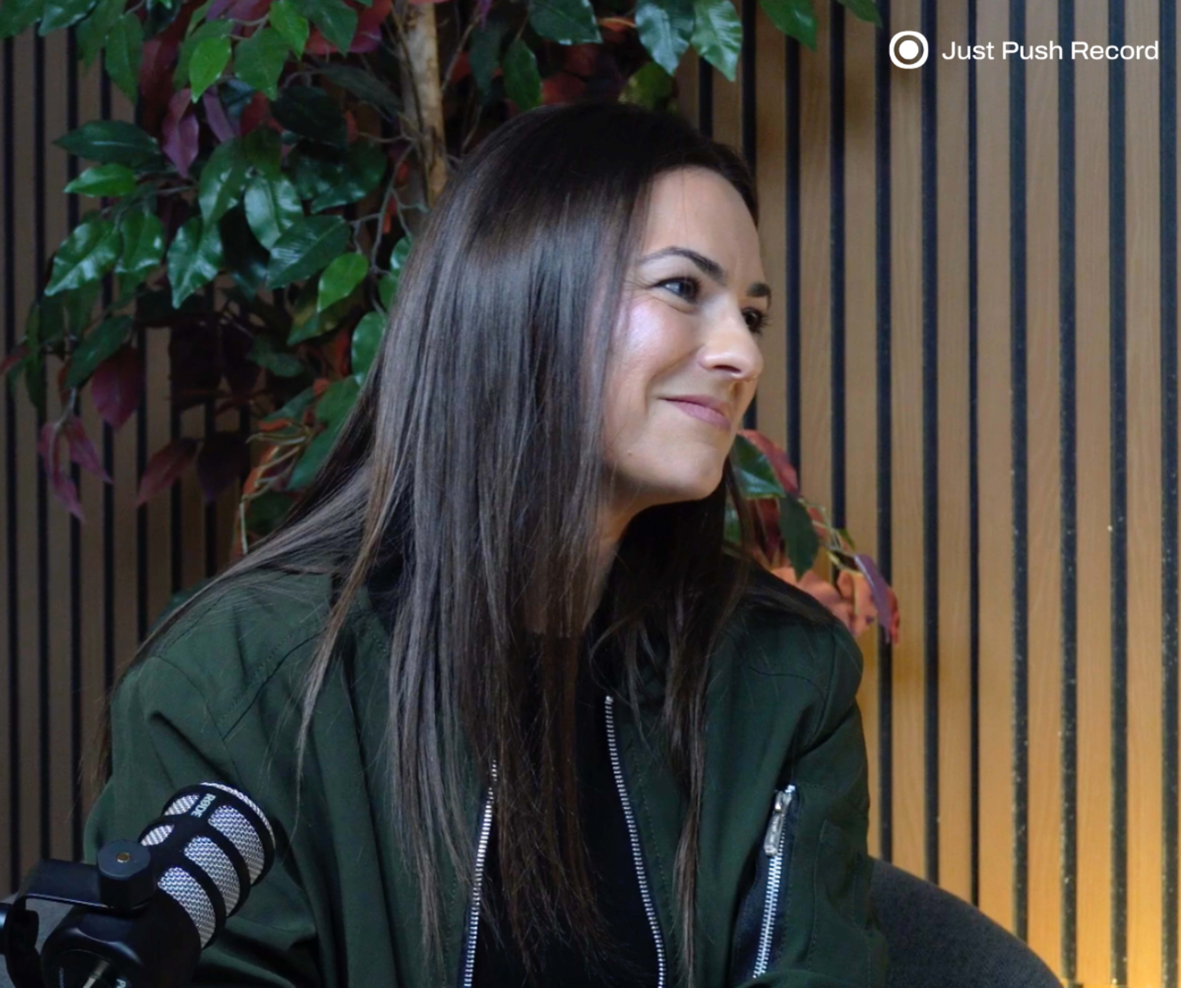 A woman with long hair sits in a recording studio, surrounded by green plants and wooden paneling.