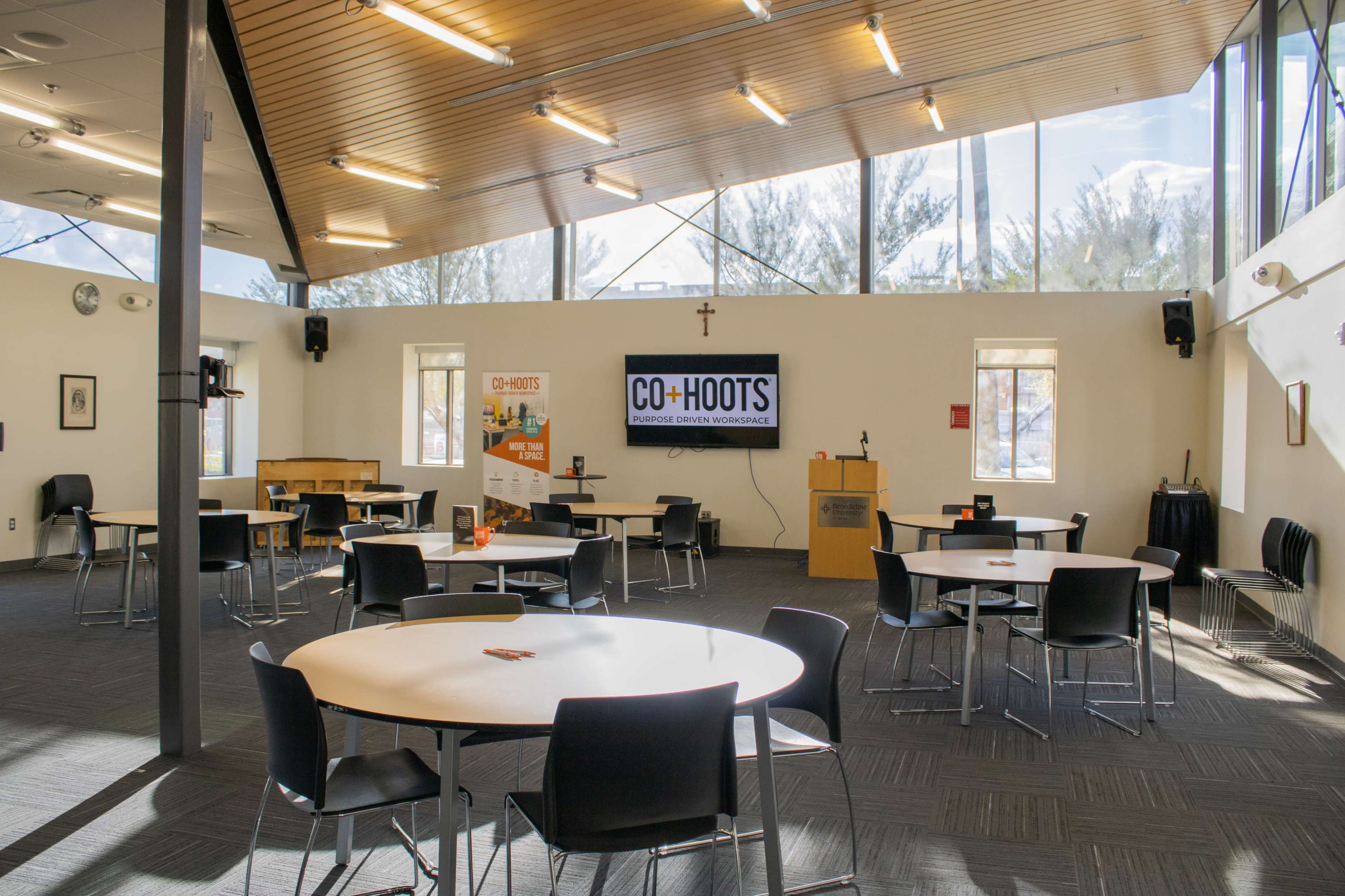 A spacious, well-lit room with several tables and chairs arranged for a meeting, featuring a large screen and a podium at one end.