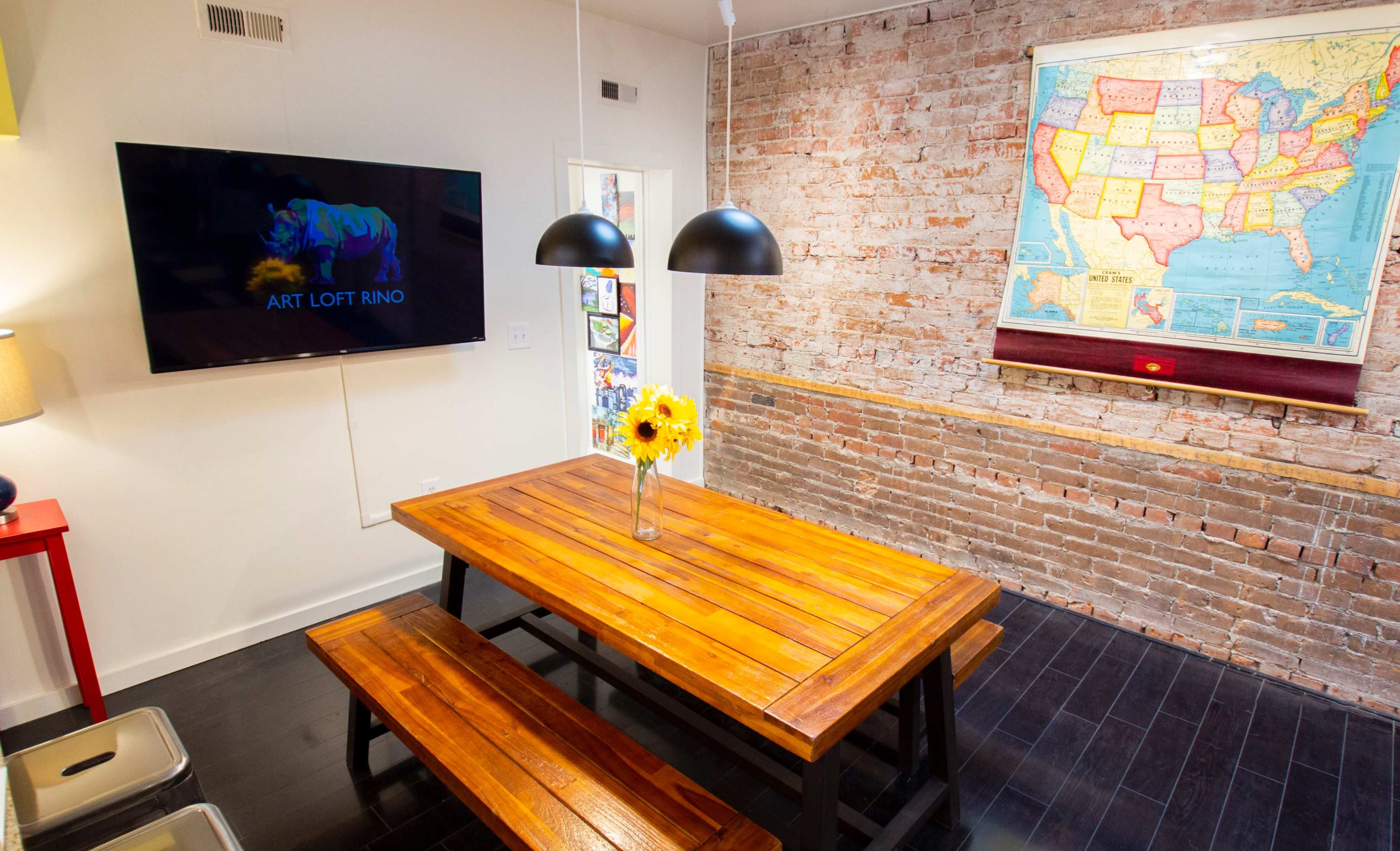 A wooden dining table with a vase of sunflowers is set under black pendant lights in a room featuring brick walls and a map of the United States.