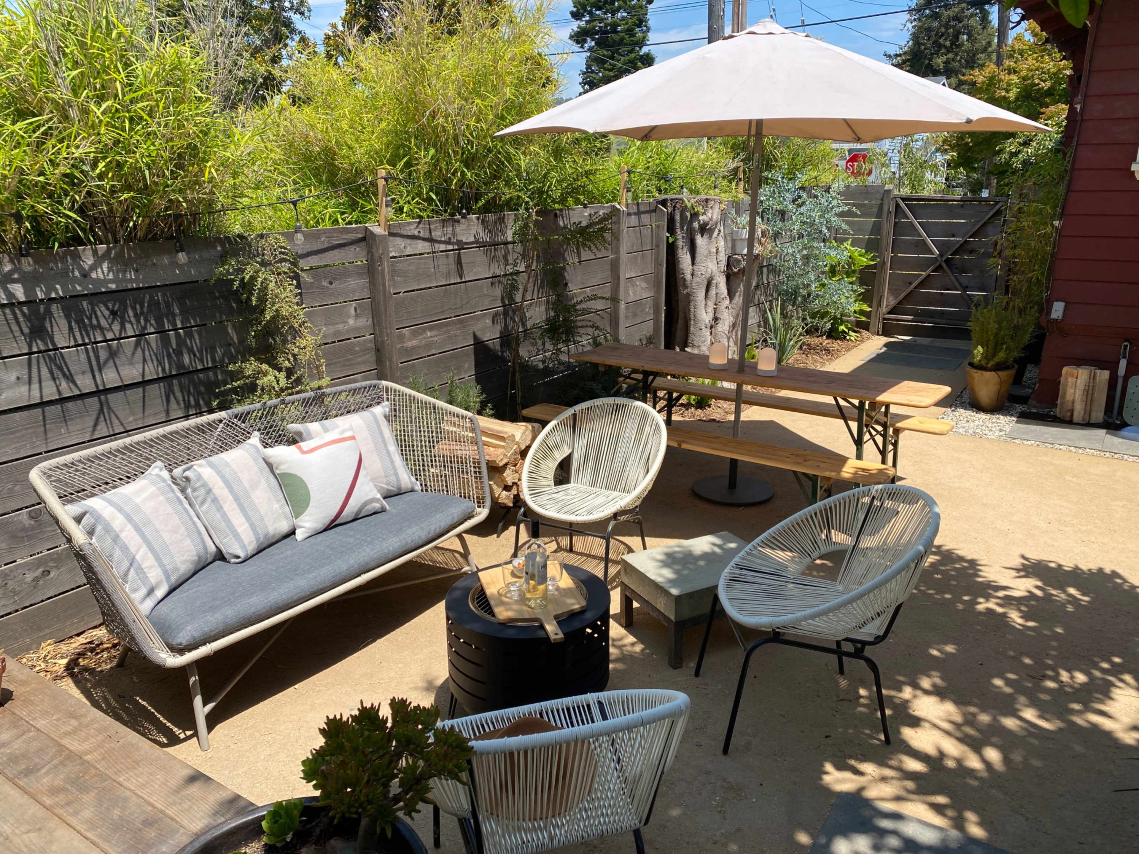 A backyard patio area with a sofa, round table, and several chairs under an umbrella, surrounded by wooden fences and greenery.