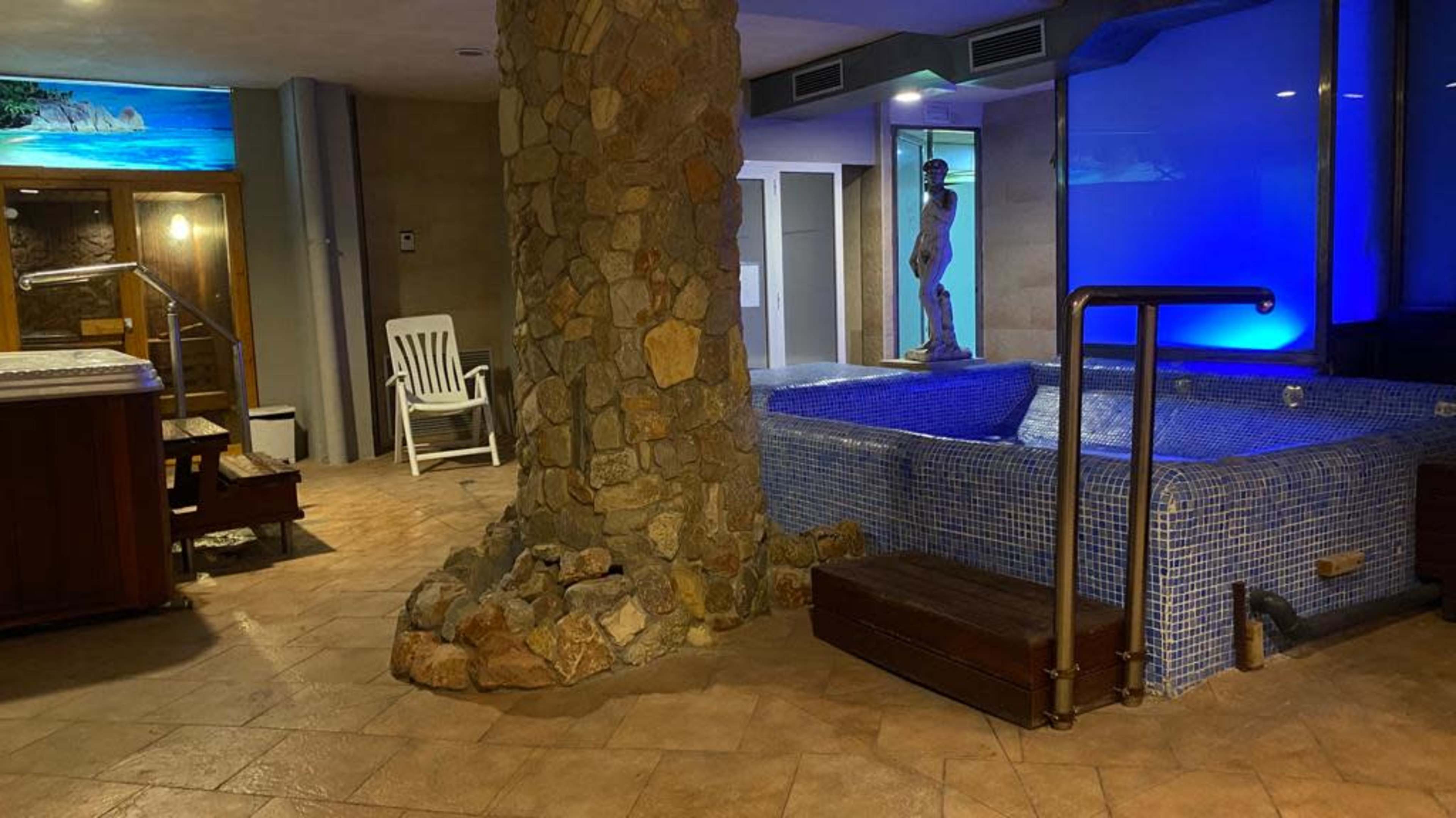 An indoor spa area featuring a hot tub, a stone pillar, a white chair, and a statue beside a blue-lit wall.