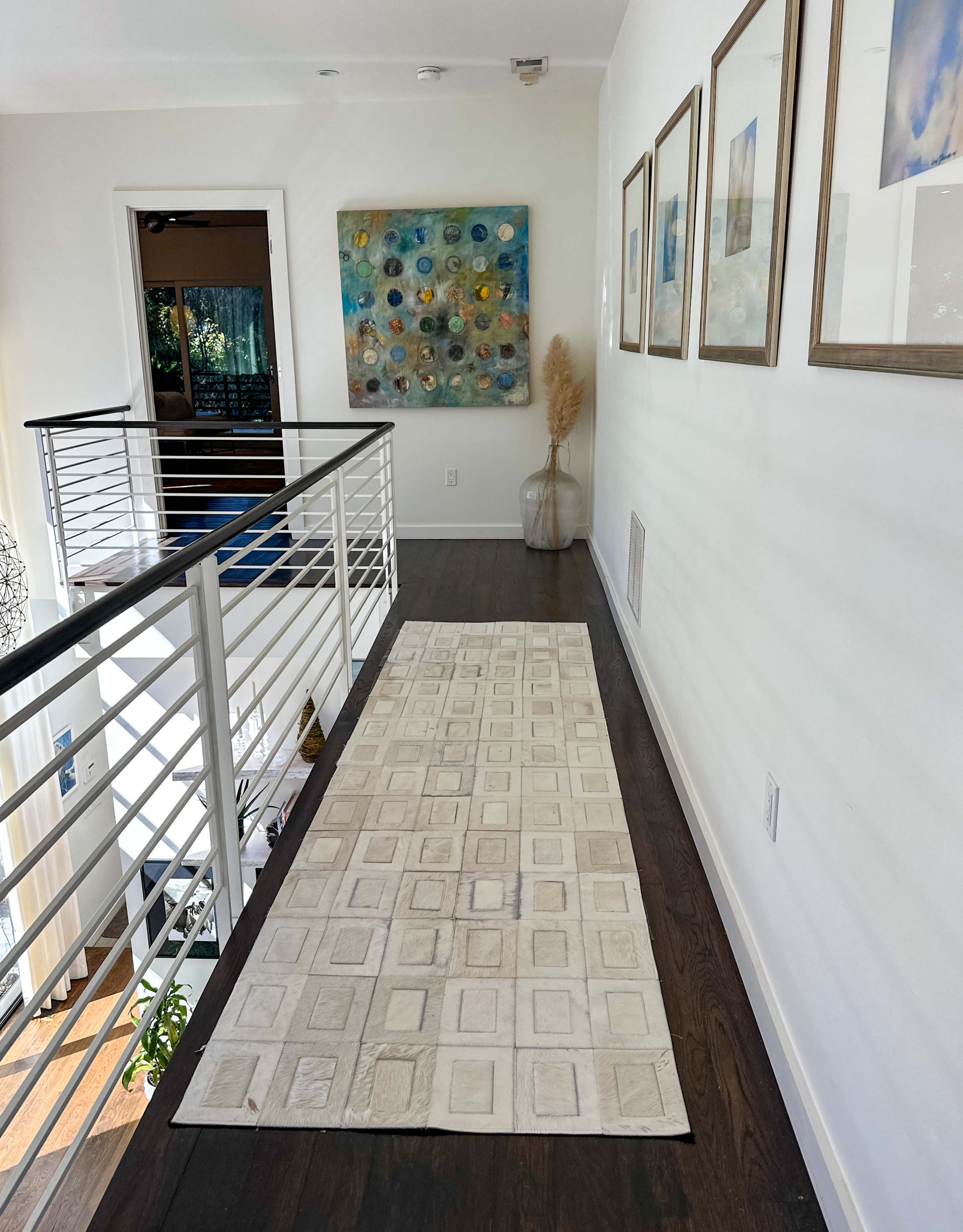 A narrow hallway features a long, rectangular rug running alongside a wall adorned with framed pictures and a colorful abstract painting at the end.