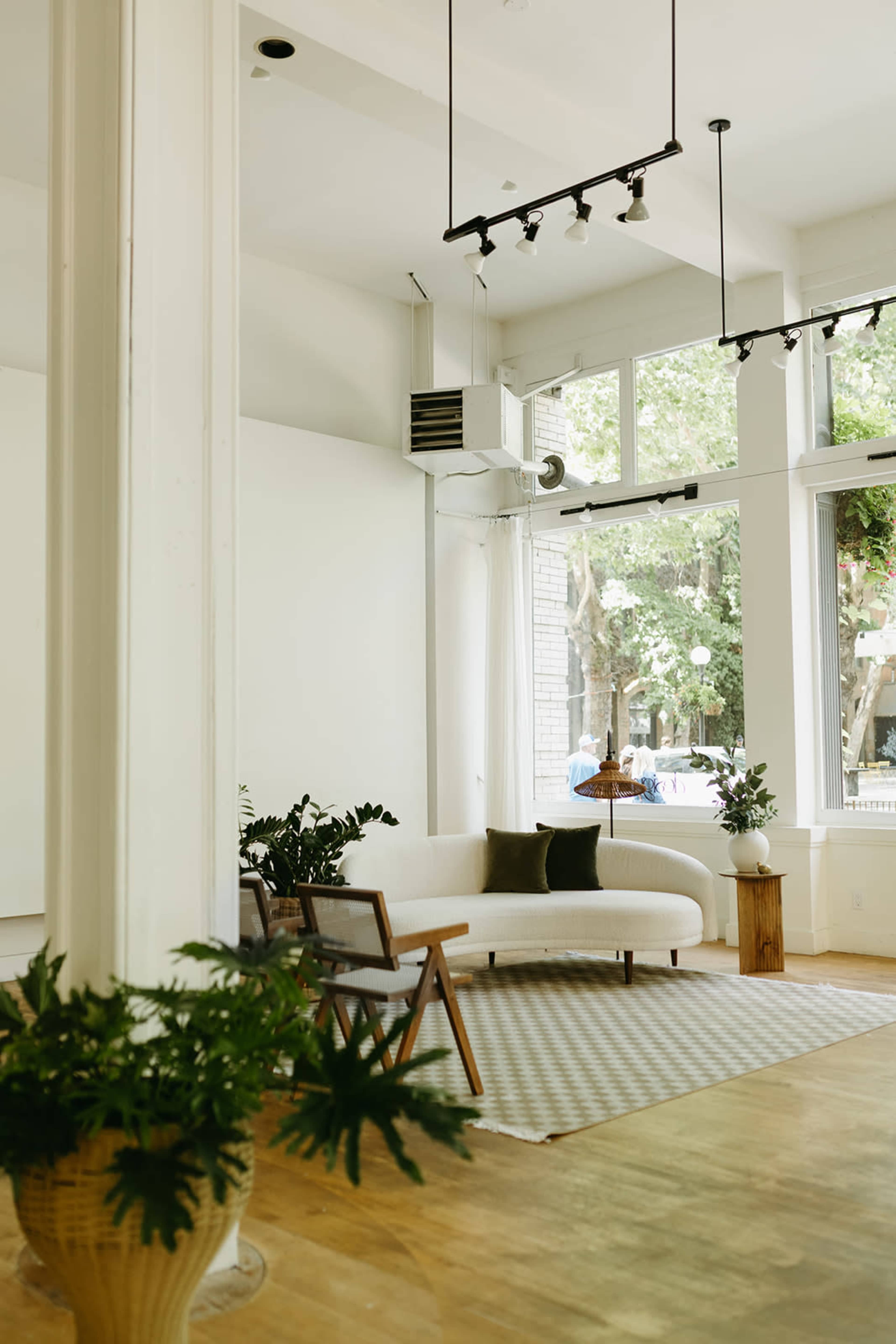 The image shows a bright and airy interior space featuring a light-colored sofa, a wooden chair, and potted plants, with large windows allowing natural light to illuminate the room.