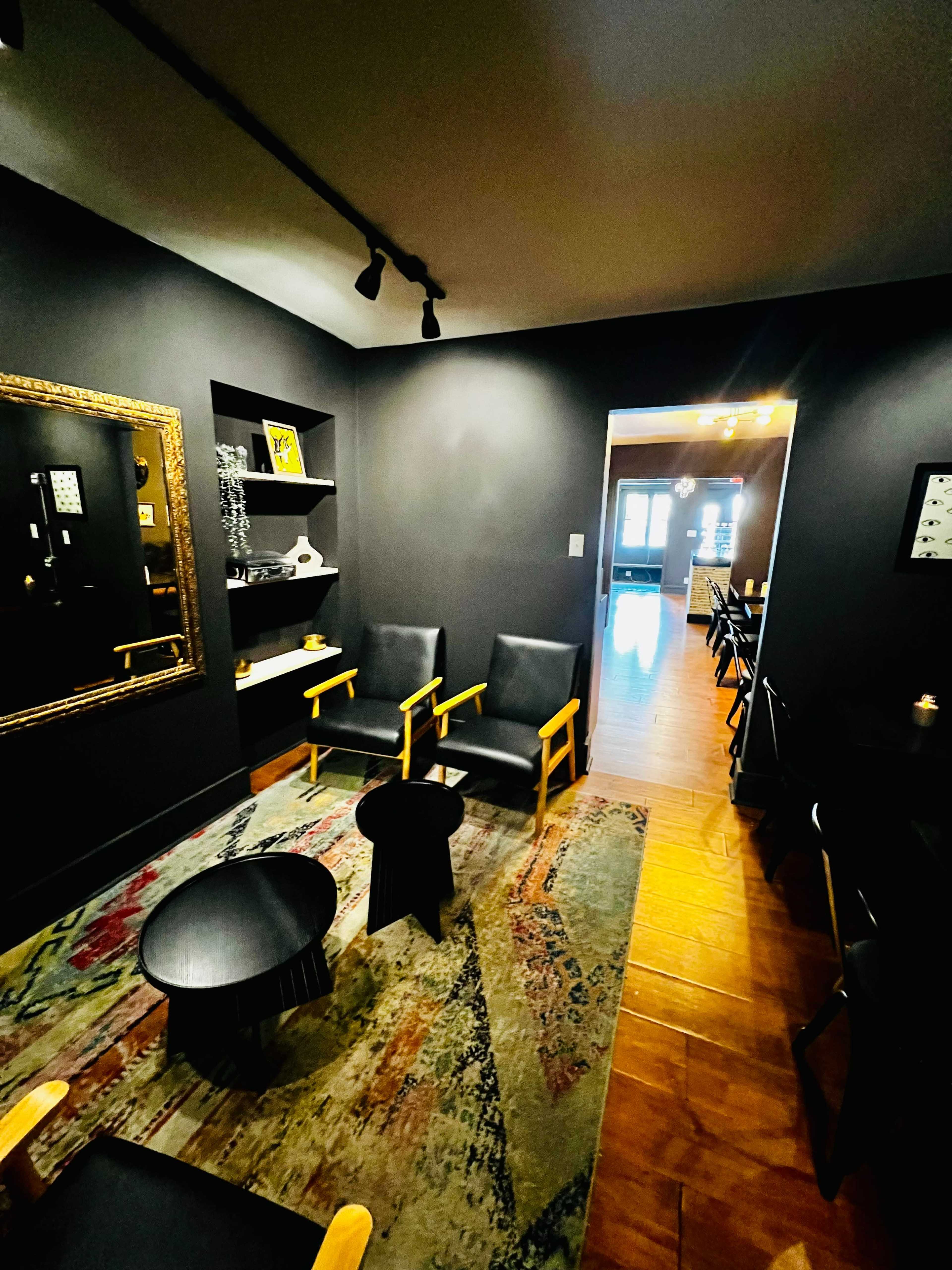 The image shows a cozy, dimly lit living area with black walls, two wooden chairs, a low table, and an entryway leading to another space.