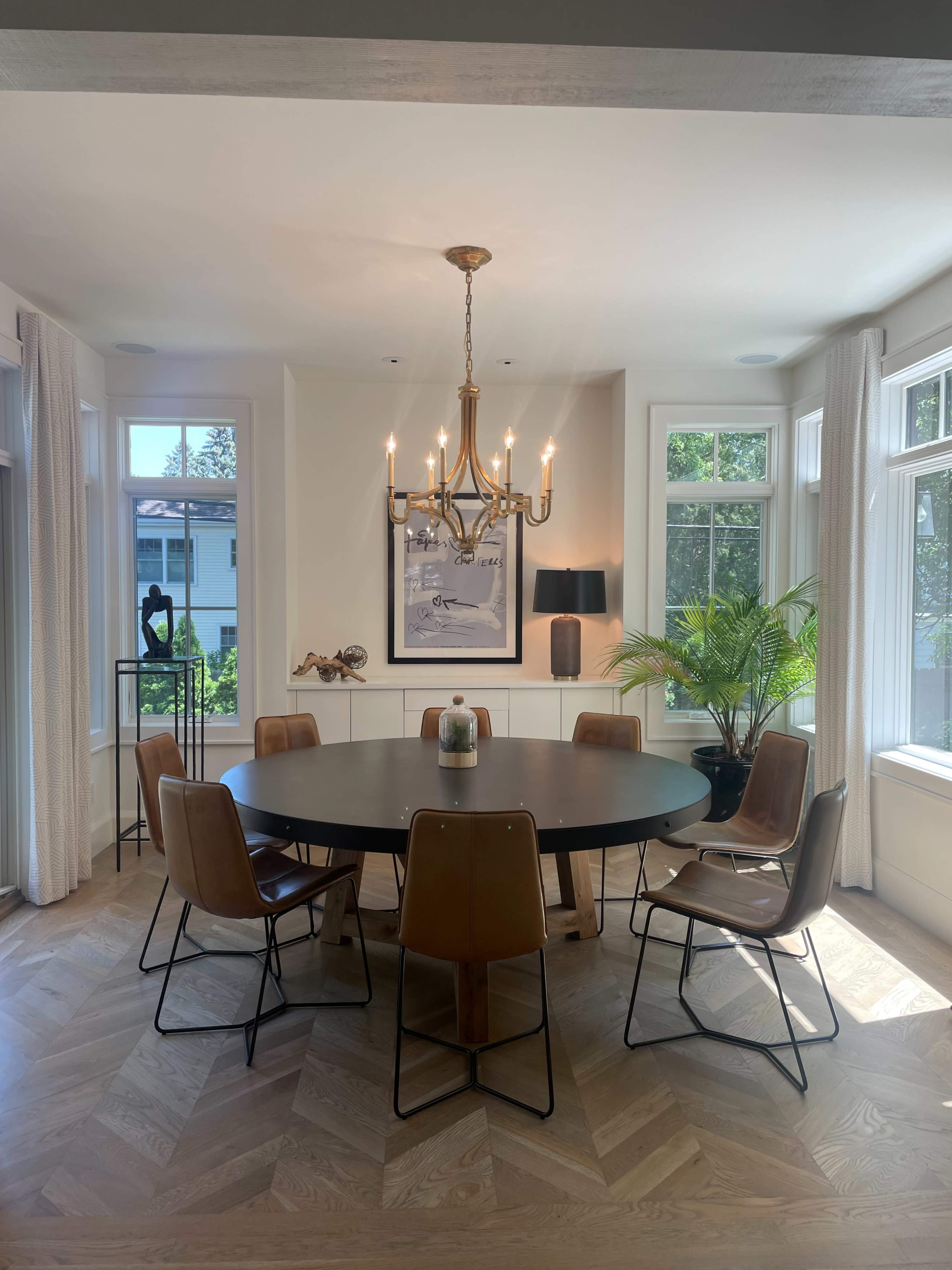 A round dining table with upholstered chairs is illuminated by a chandelier in a well-lit dining room featuring large windows and decorative plants.