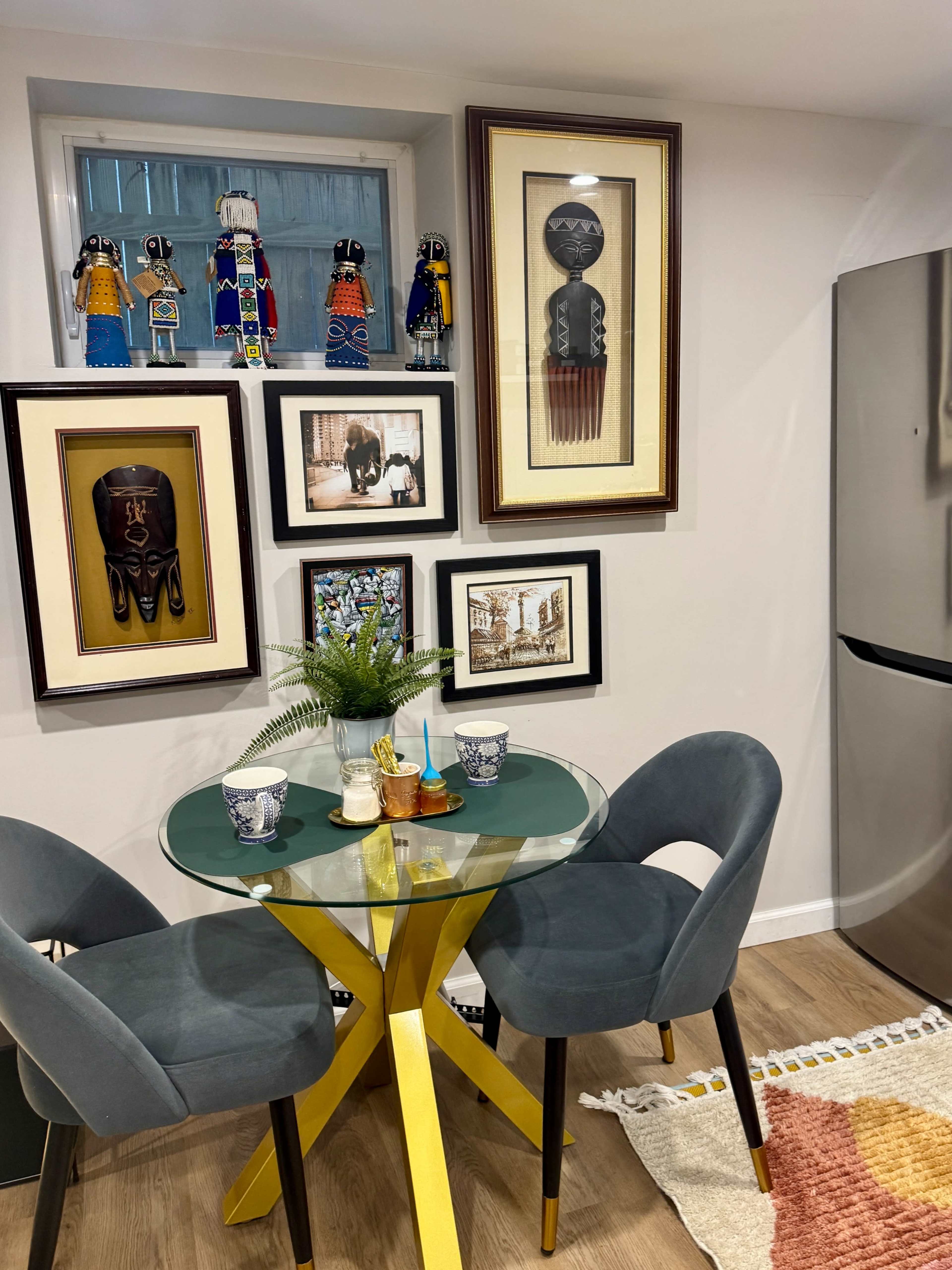 A small dining area features a round glass table with yellow legs, two gray chairs, and a collection of framed artwork arranged on the wall.