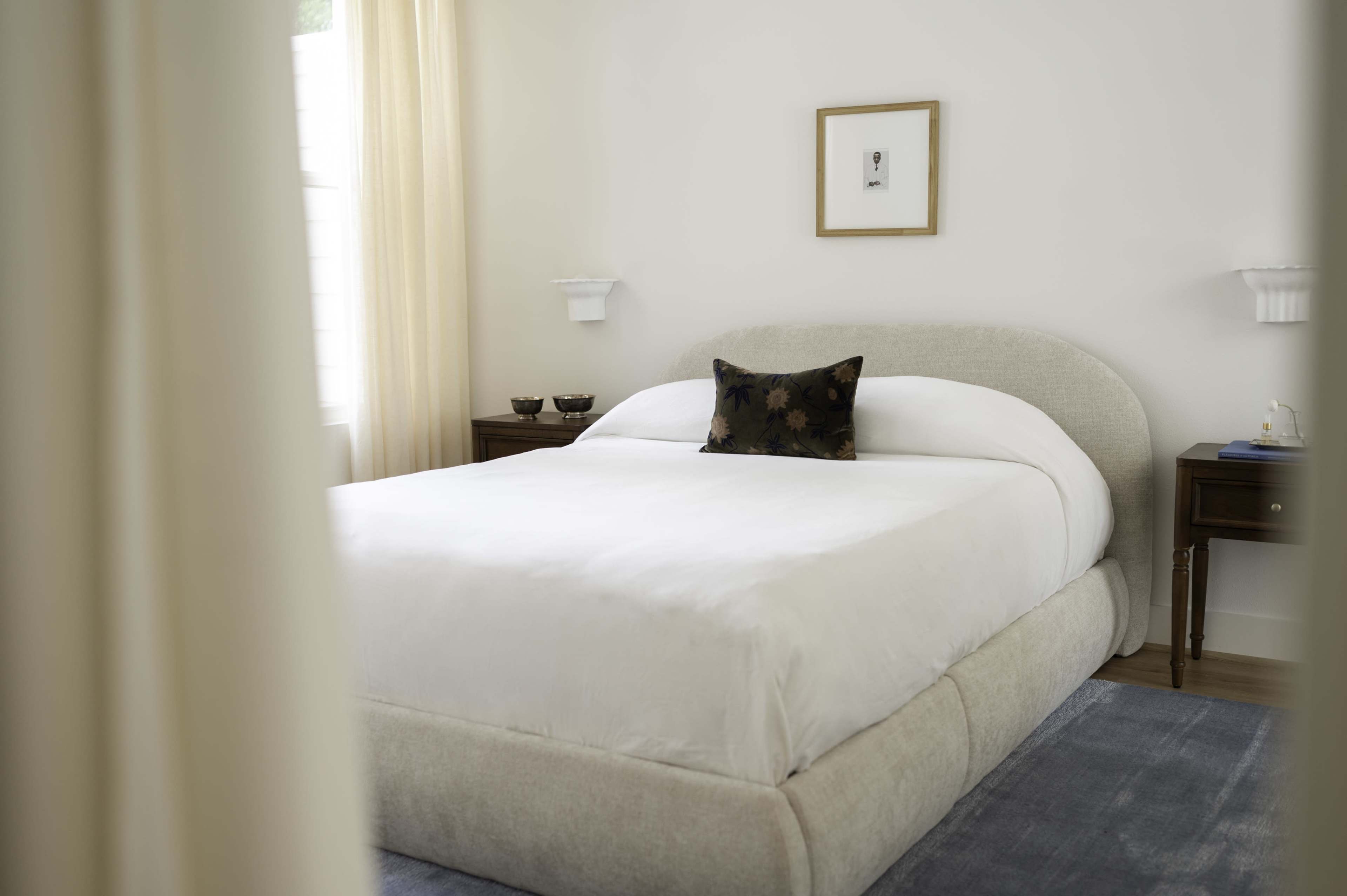 The image shows a neatly made bed with a decorative pillow in a well-lit bedroom featuring soft curtains and minimalistic furniture.