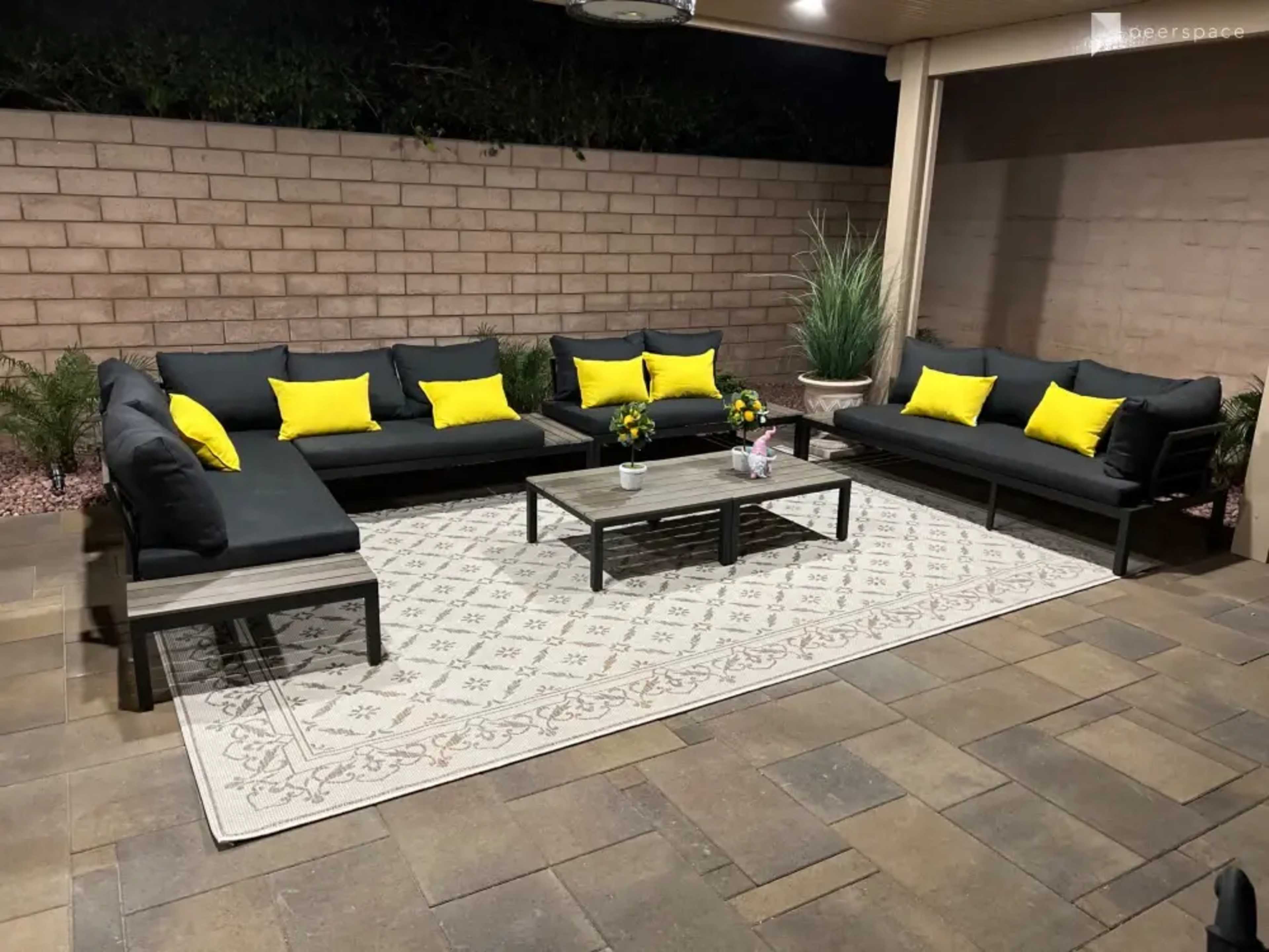 A patio area features a black sectional sofa with yellow cushions, a central table, and a large area rug on stone flooring.