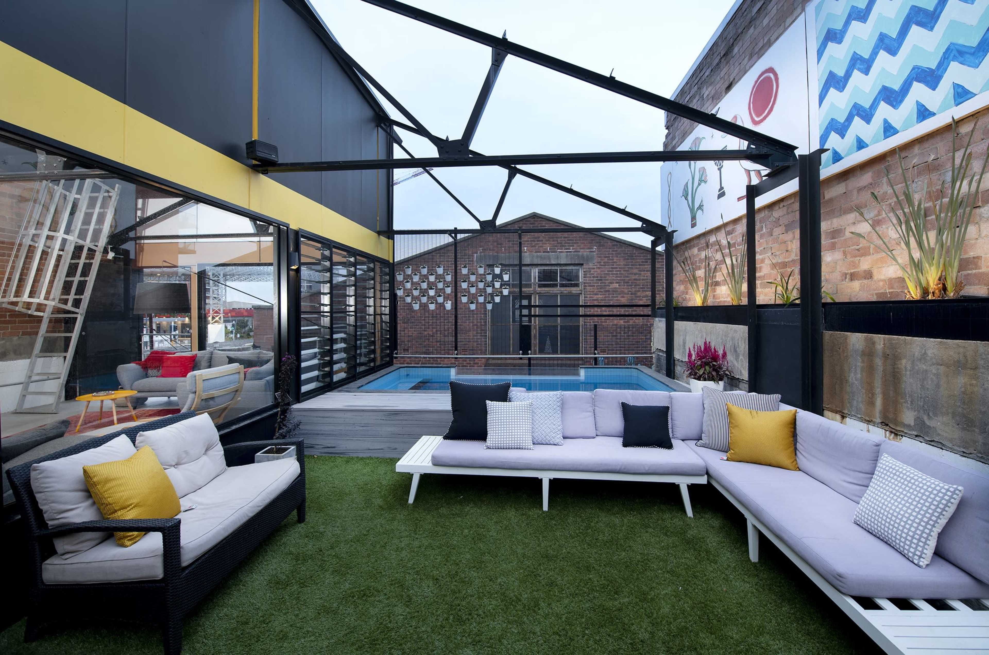 A modern outdoor lounge area with a swimming pool, featuring a sectional sofa with yellow and gray cushions and a backdrop of a brick wall and colorful murals.