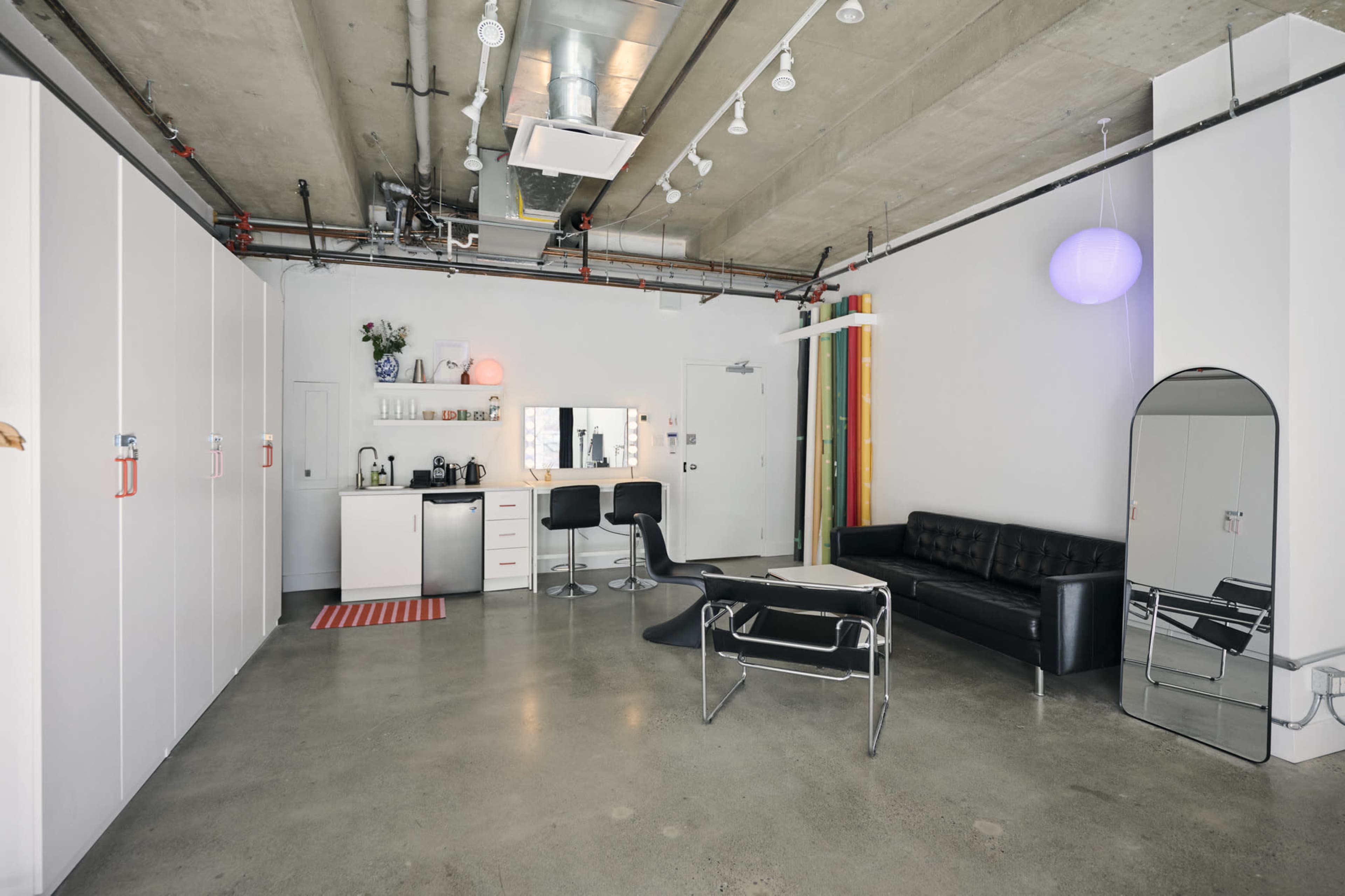 The image shows an industrial-style apartment featuring a black leather couch, a kitchenette with white cabinets, and a minimalist seating area.