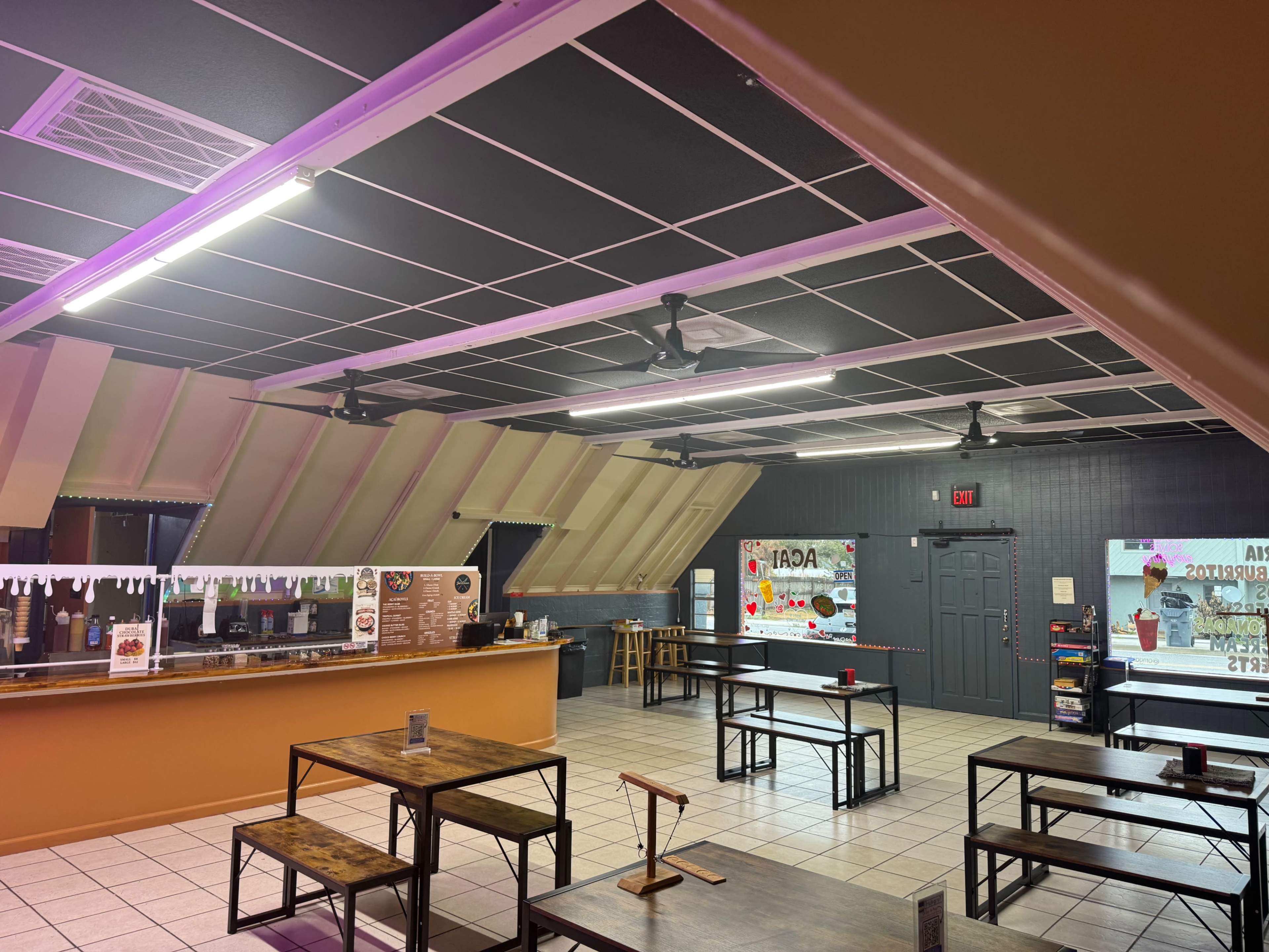 The image shows an indoor café with several wooden tables, ceiling fans, and a service area displaying a menu on the wall.