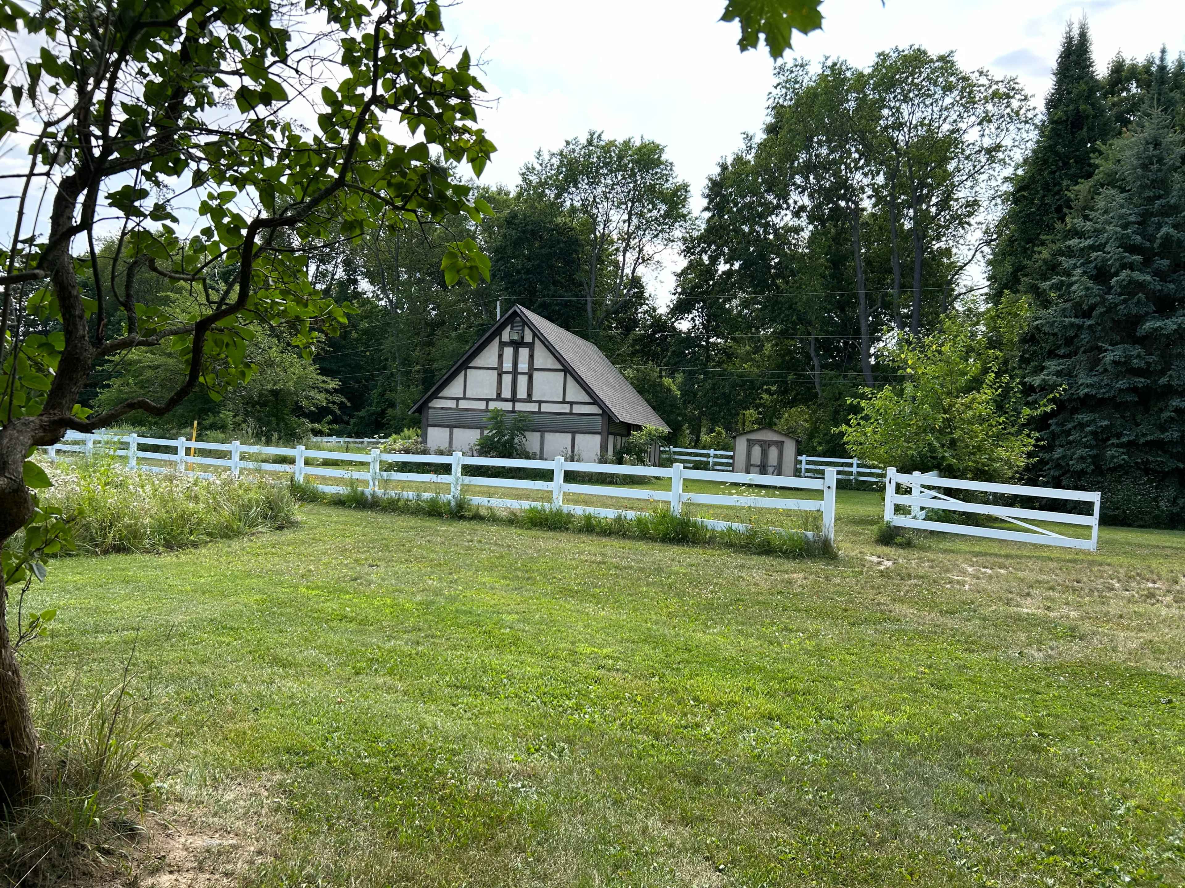 A small, cottage-style house with a sloped roof is situated behind a white picket fence, surrounded by green grass and trees.