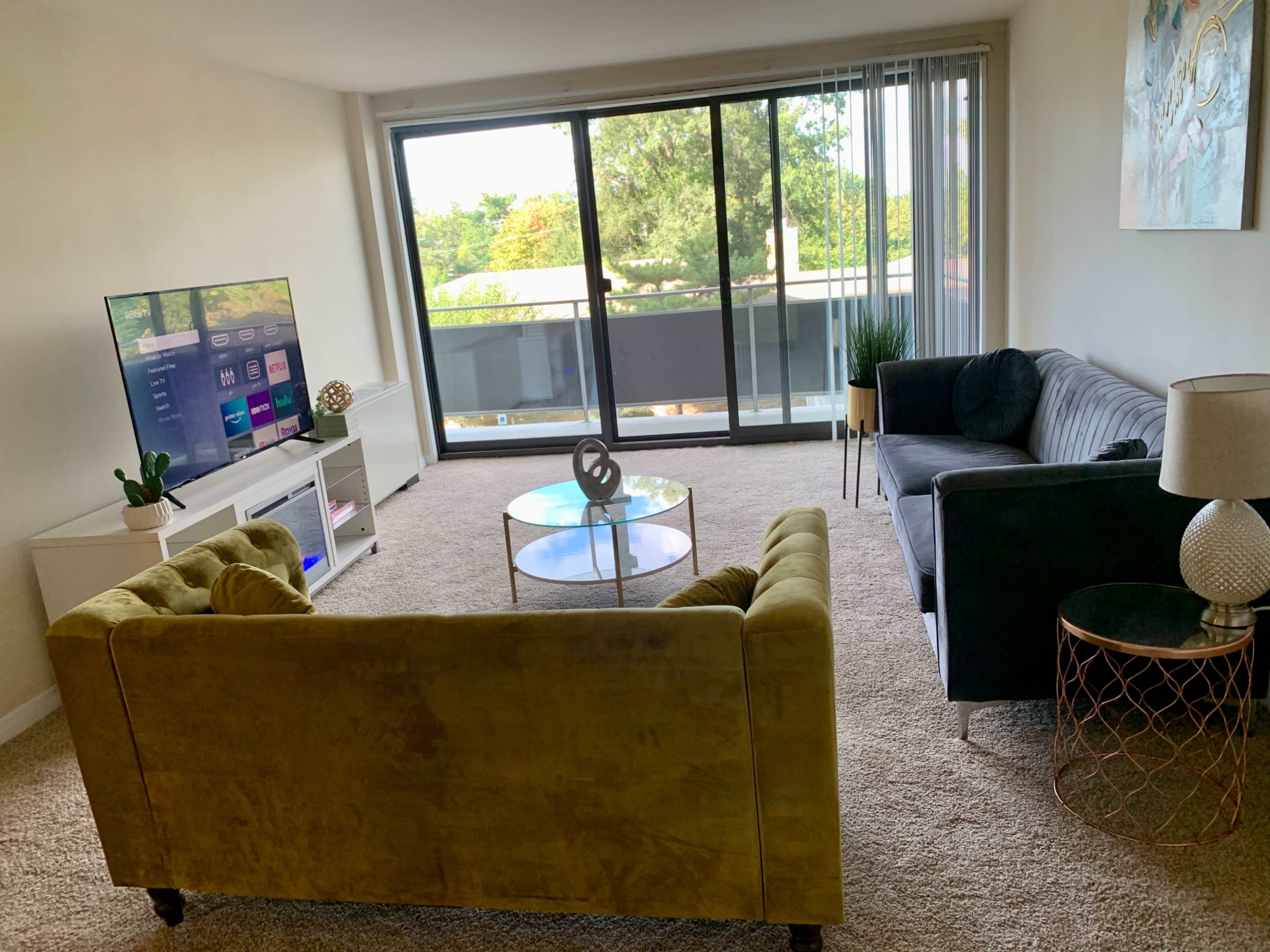 A bright living room features a yellow sofa and a dark gray couch facing a glass coffee table, with a large TV and sliding glass door leading to a balcony.