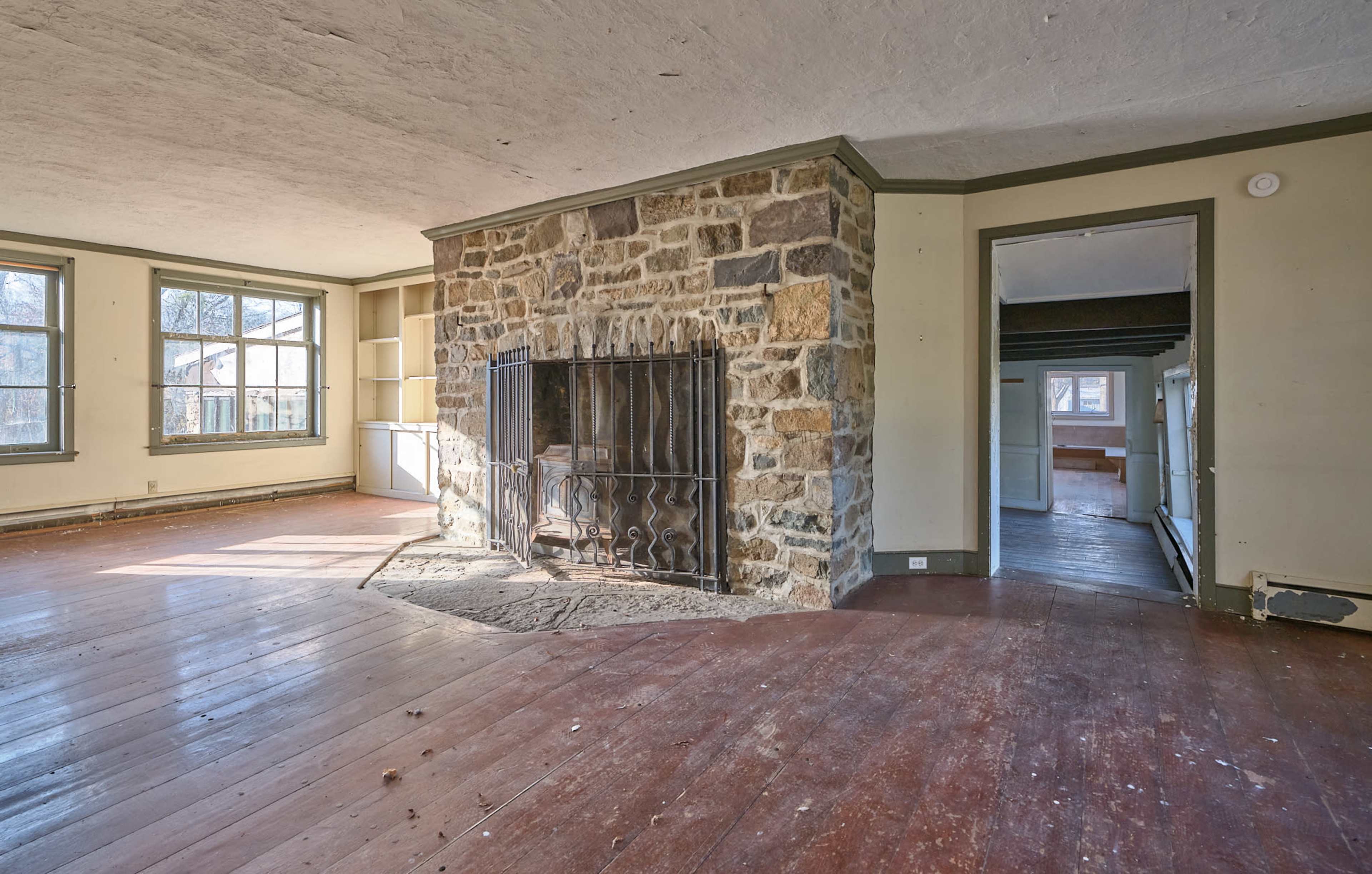 The image shows an empty room with a stone fireplace as the focal point, featuring large windows and hardwood flooring.