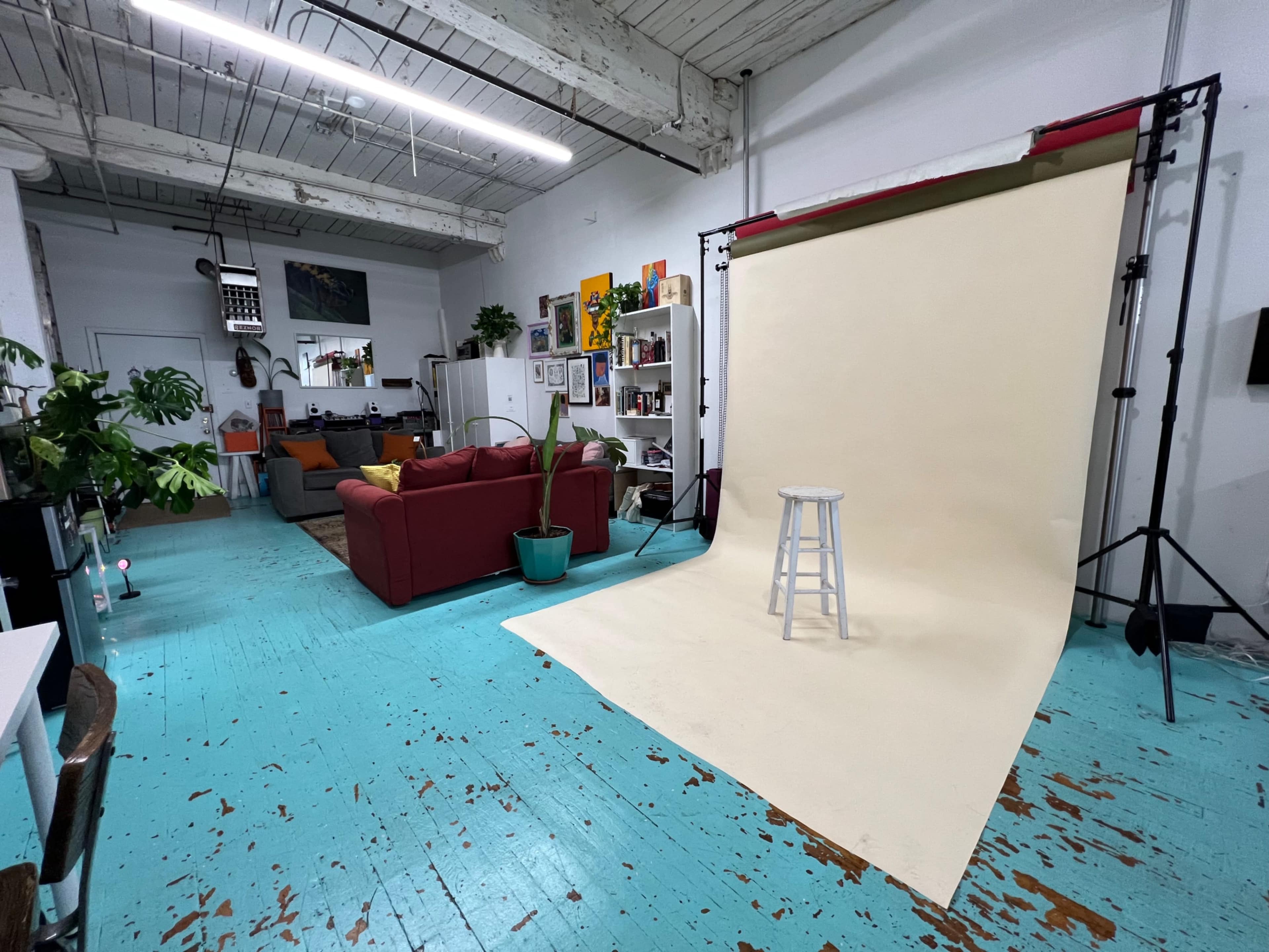 A spacious studio with a beige backdrop on a stand, a white stool in front of it, a red sofa, and various plants and shelves in the background.