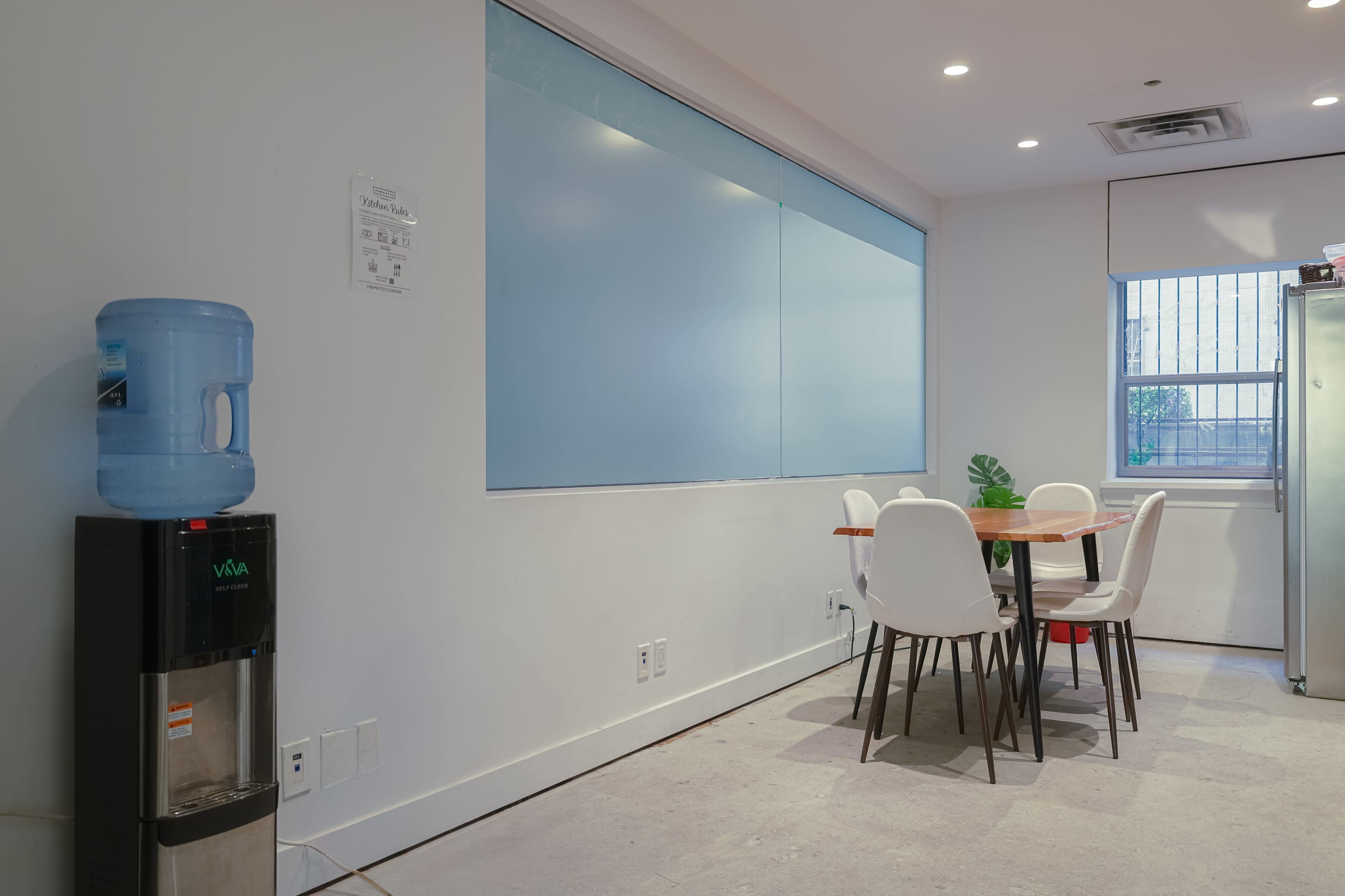 The image shows a small kitchen area featuring a water cooler, a round dining table with white chairs, and a large window with frosted glass.