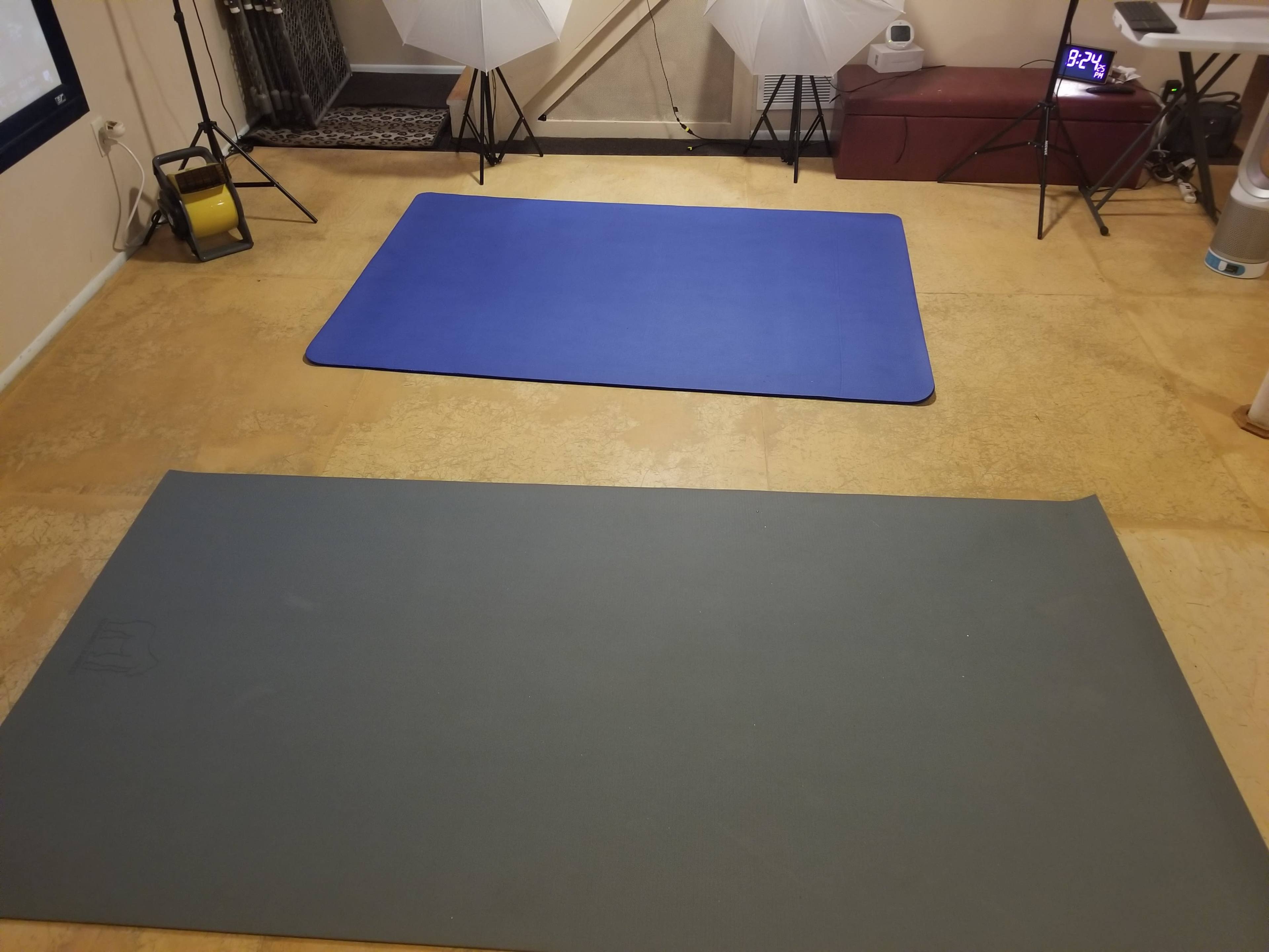 The image shows two exercise mats on a textured floor, with a clock and lighting equipment in the background.