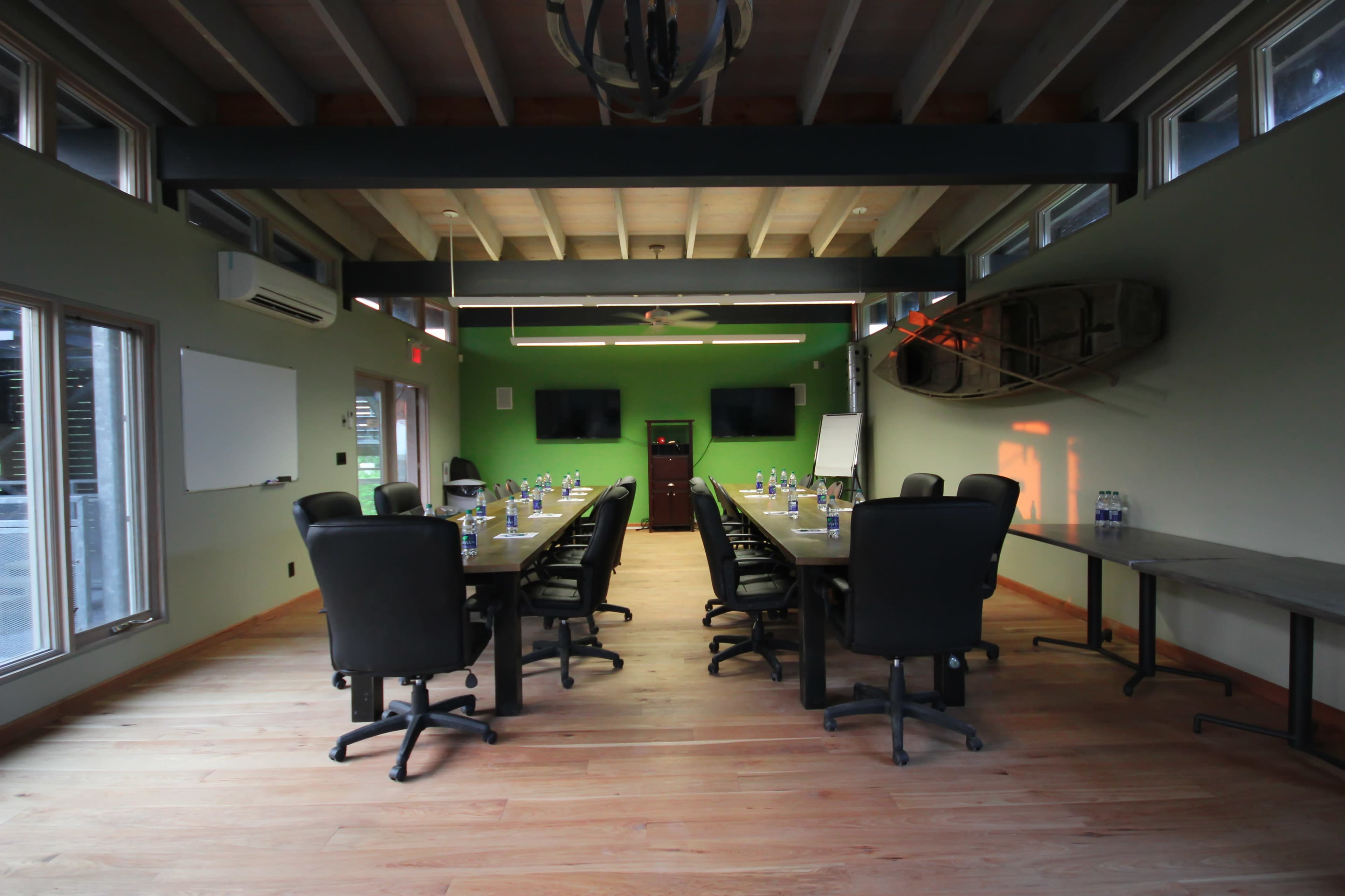 A conference room with a long table surrounded by black chairs, equipped with two wall-mounted screens and large windows, features a green accent wall and wooden floors.