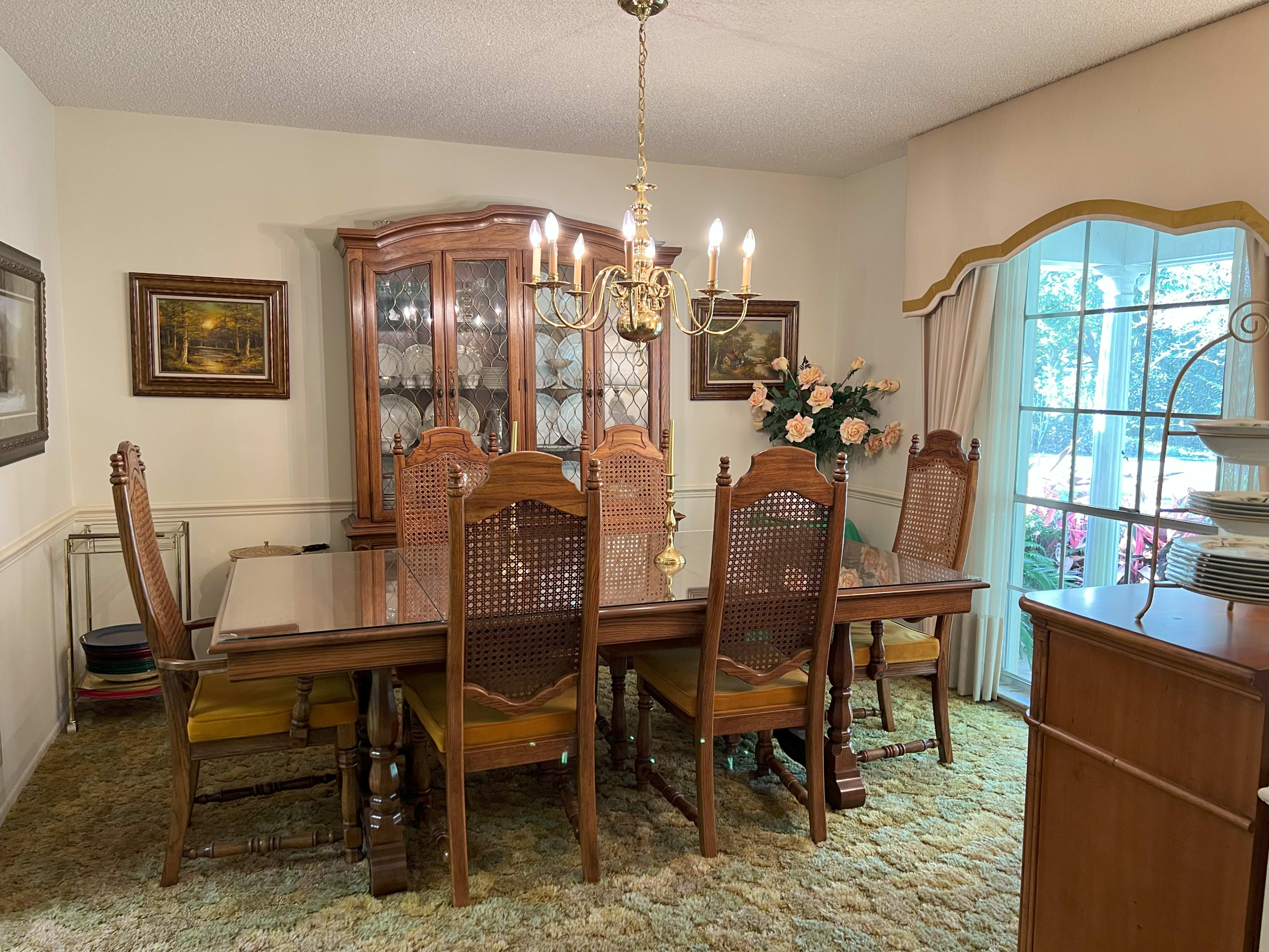 The image shows a dining room with a large wooden table surrounded by six chairs, a china cabinet filled with dishes, and a window adorned with curtains beside a floral arrangement.