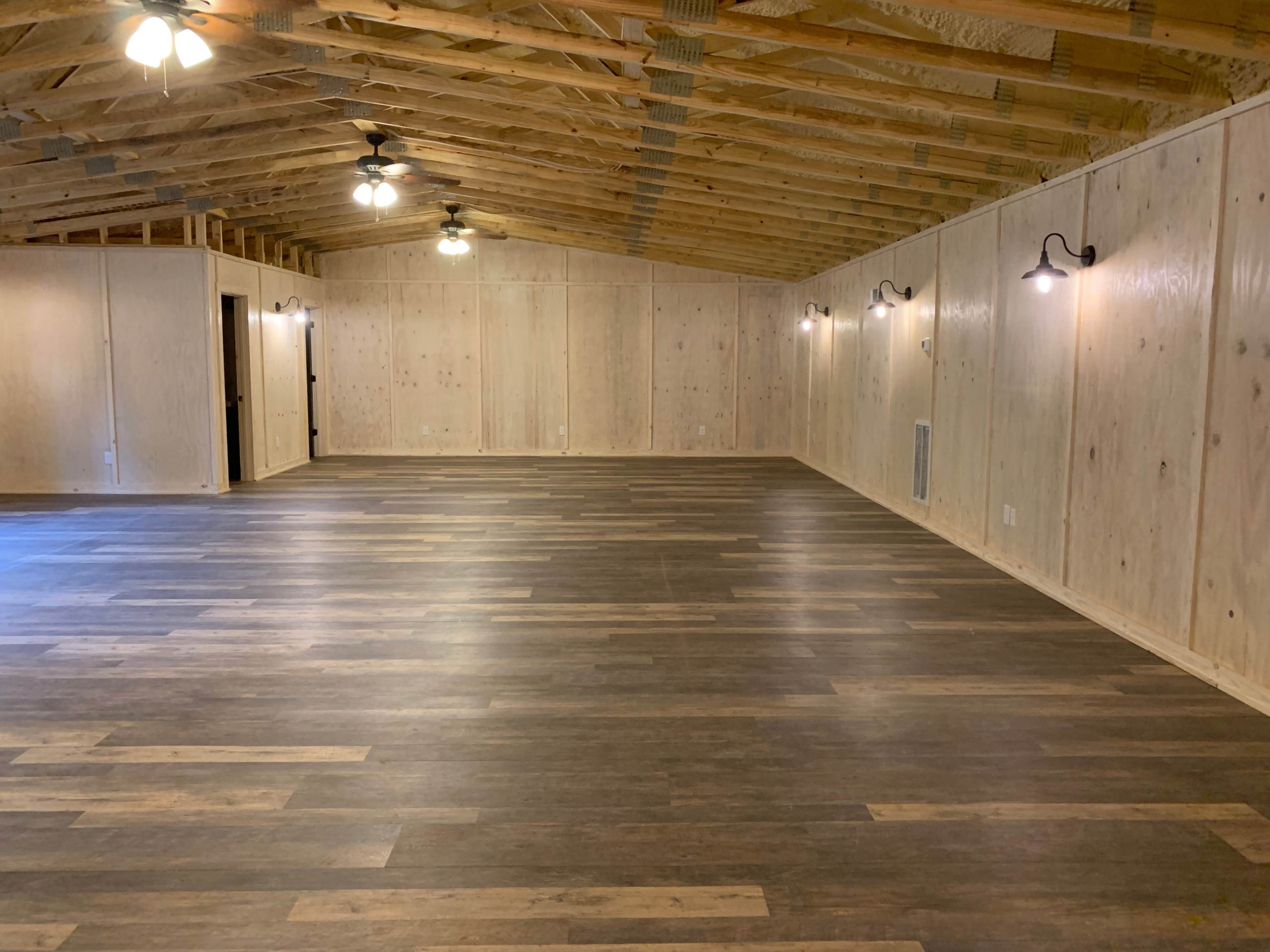 A spacious, unfinished interior room with wooden walls, a wood-beamed ceiling, and laminate flooring, illuminated by wall-mounted lights.