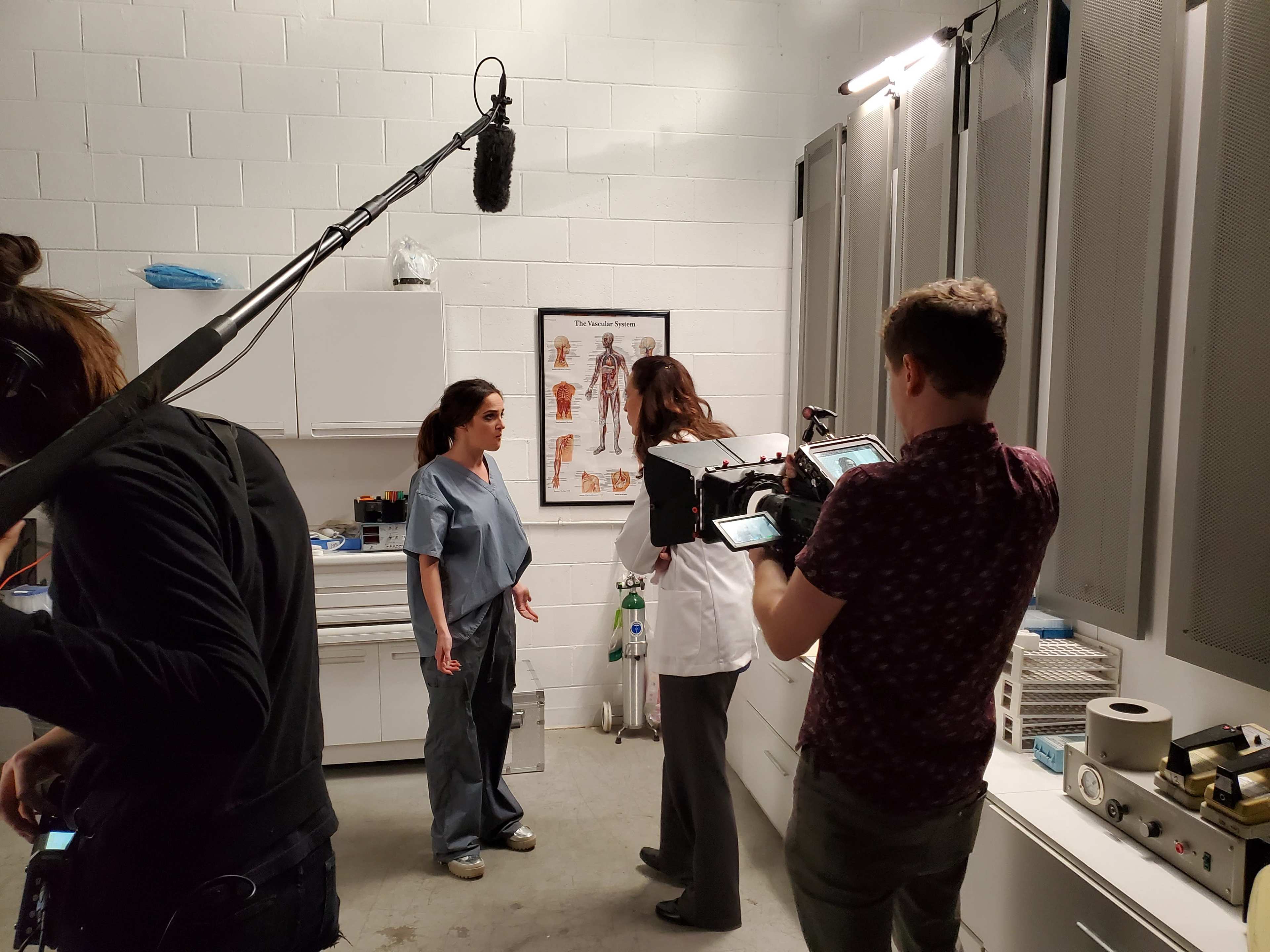 A small medical room is shown, with two women in scrubs conversing while two men operate filming equipment.