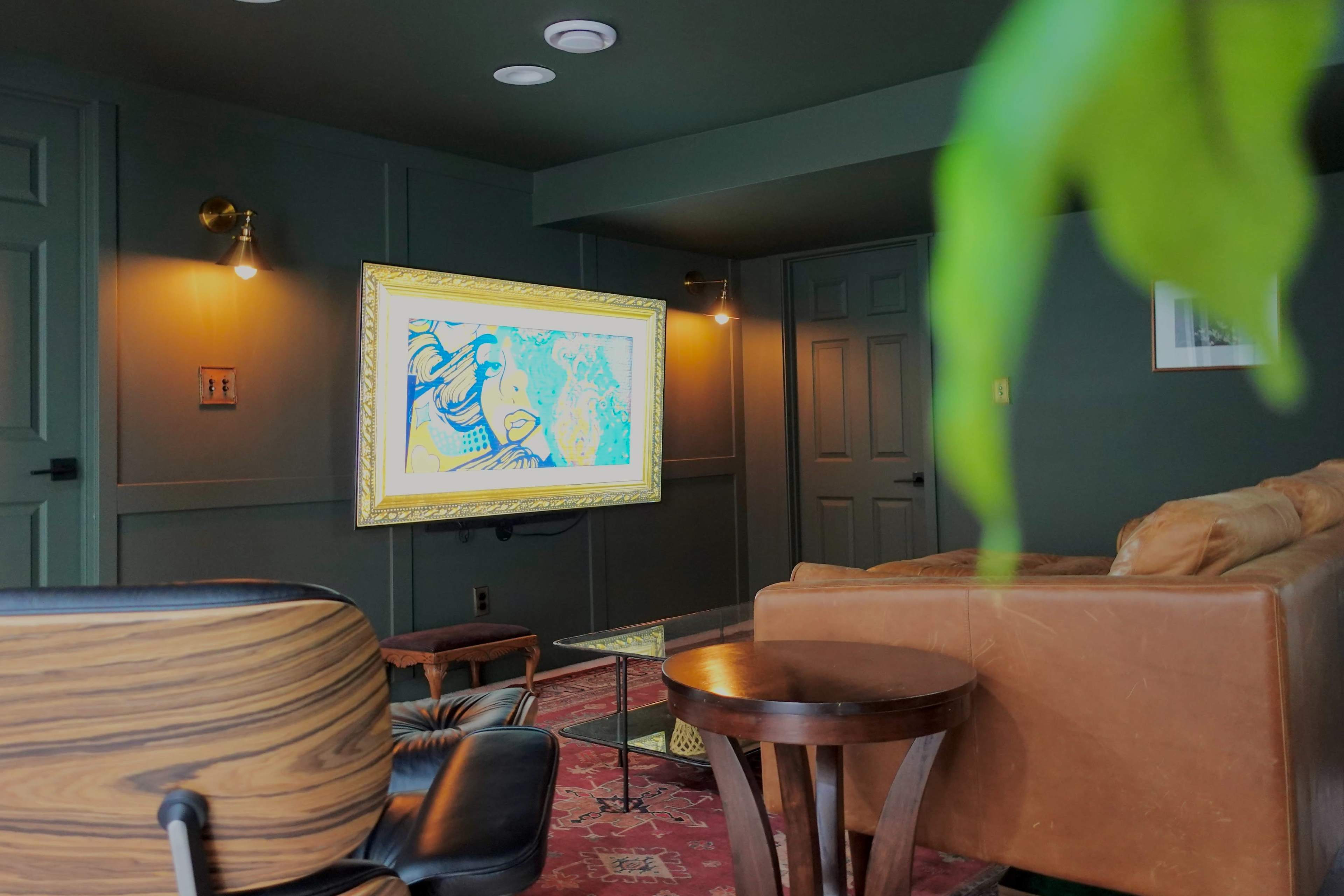 A living room features a framed artwork displayed on a wall-mounted screen, with a leather sofa and wooden furniture in the foreground.