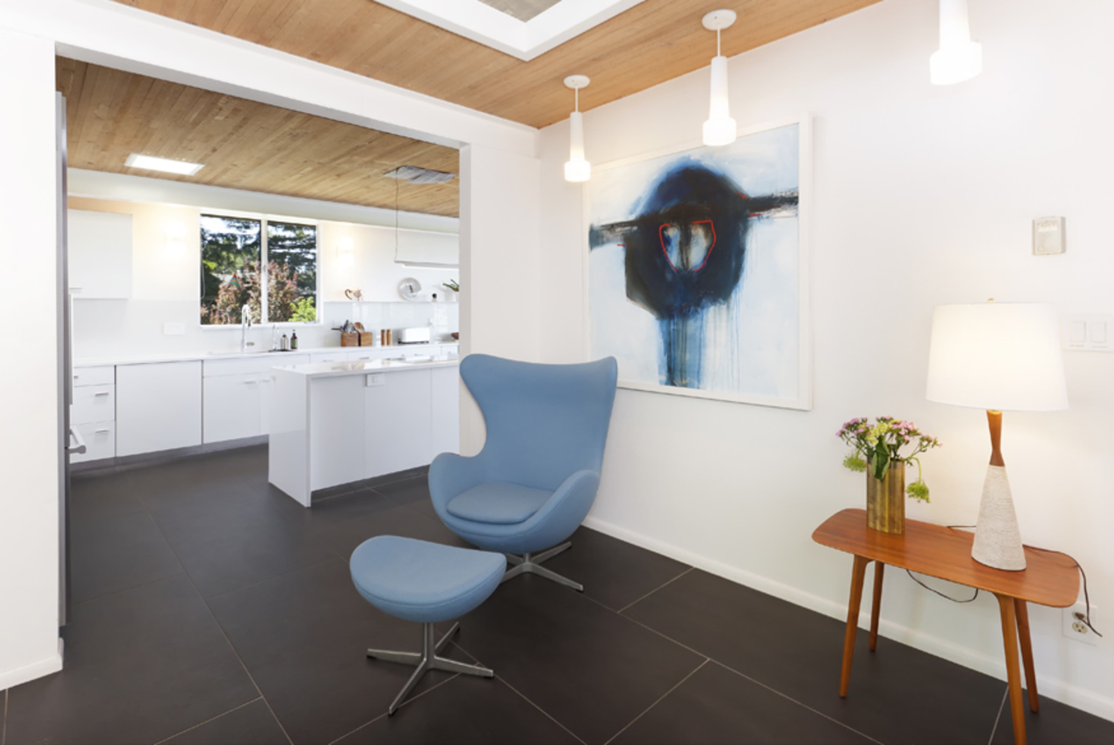 A modern living space with a blue chair and a small wooden table next to a lamp, leading into a bright kitchen area.