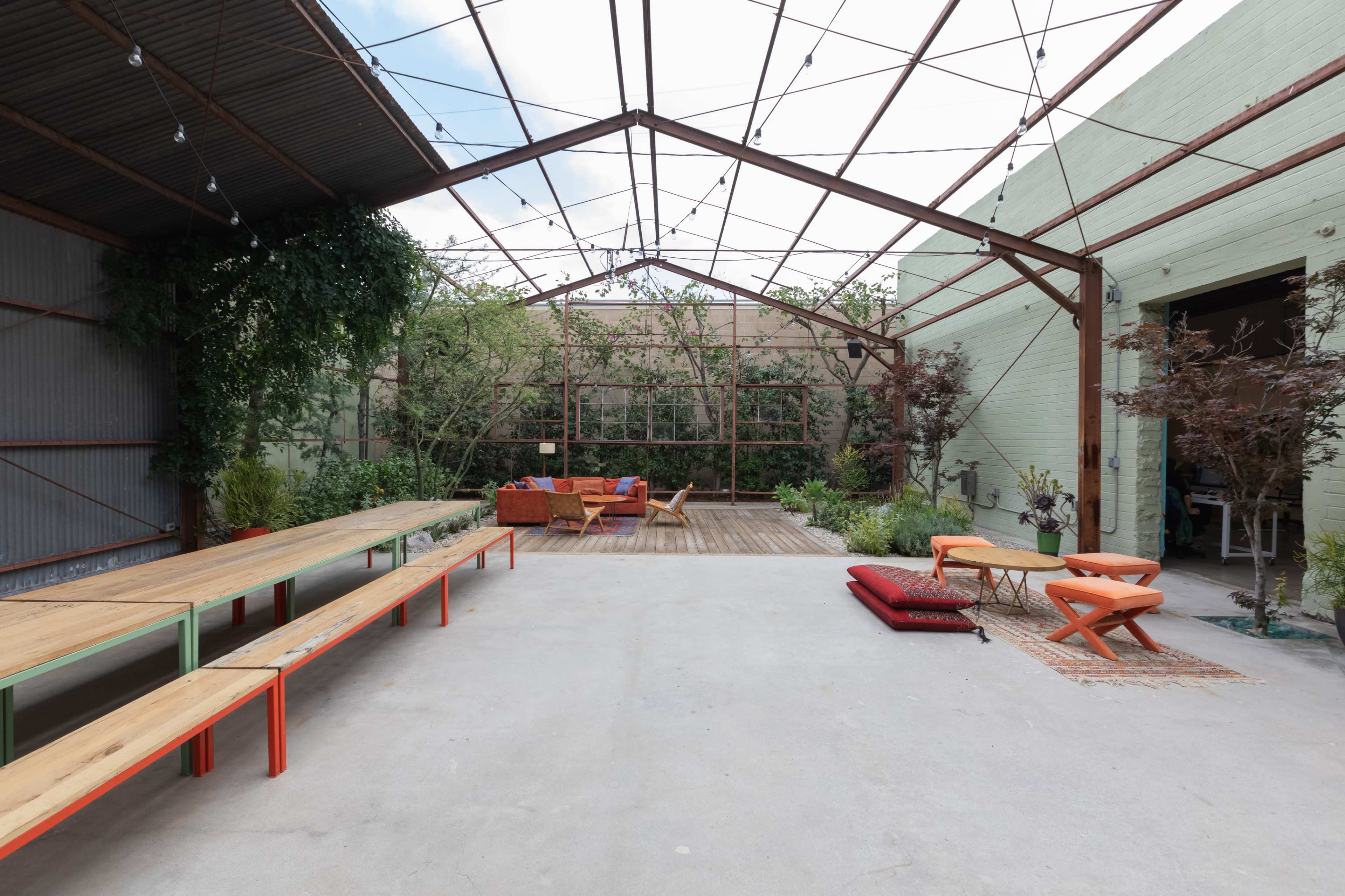 An open, industrial-style space with wooden benches, colorful seating arrangements, and greenery surrounding a wooden deck area.