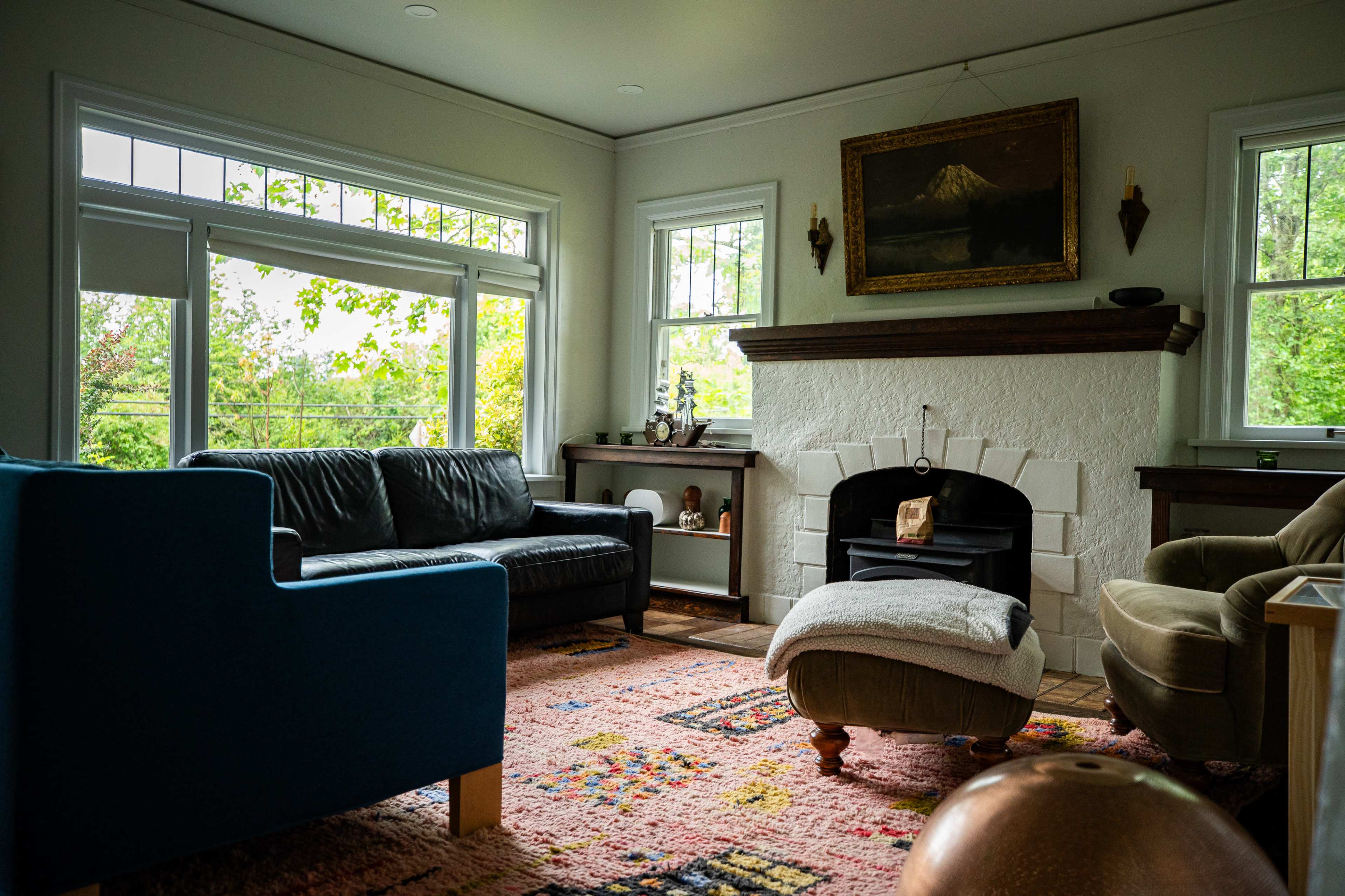 The living room features a seating area with a dark leather sofa, a blue upholstered chair, a green ottoman, and a white fireplace adorned with a painting and natural light coming through the windows.