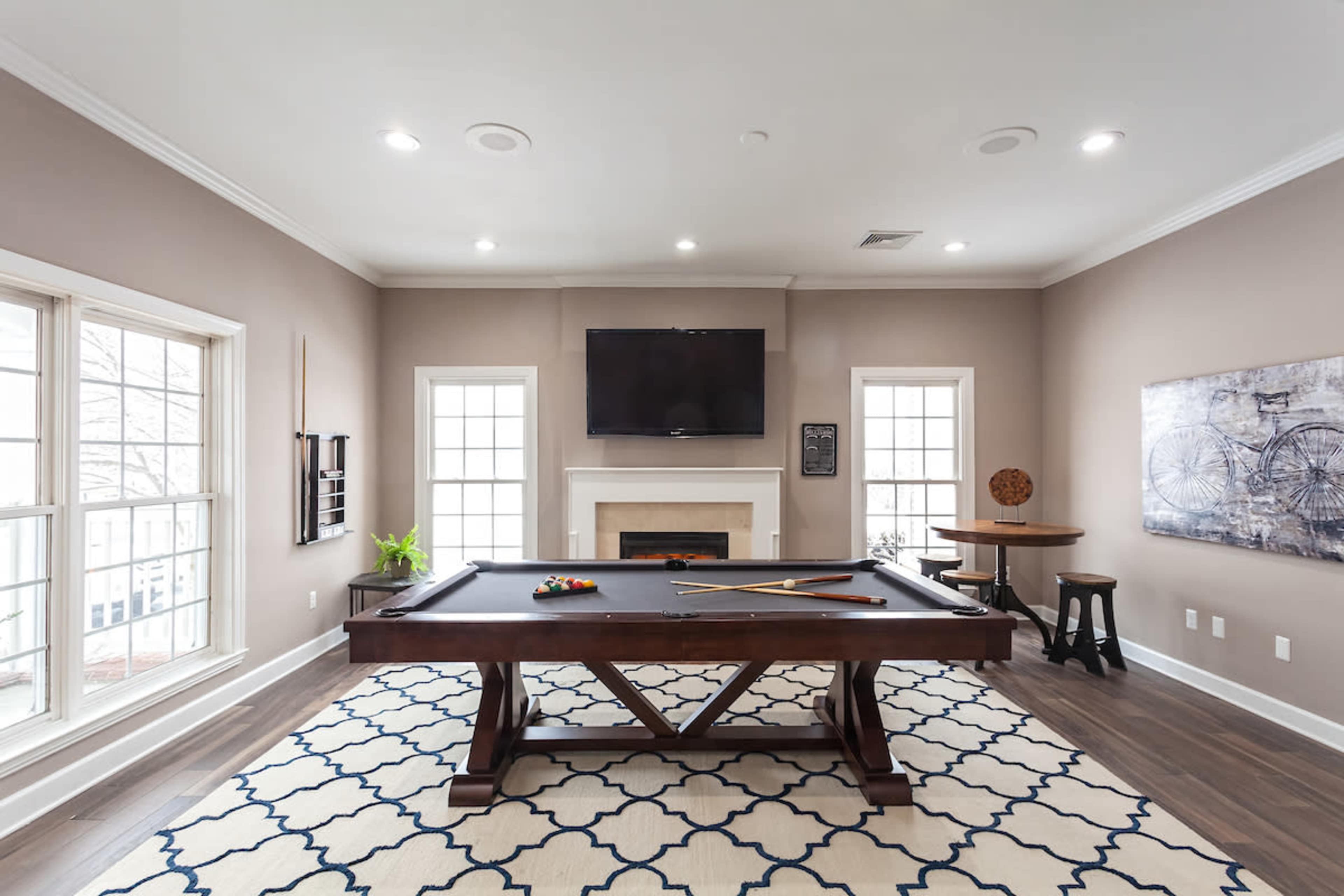The image shows a game room featuring a billiard table, a television mounted on the wall, and large windows allowing natural light into the space.