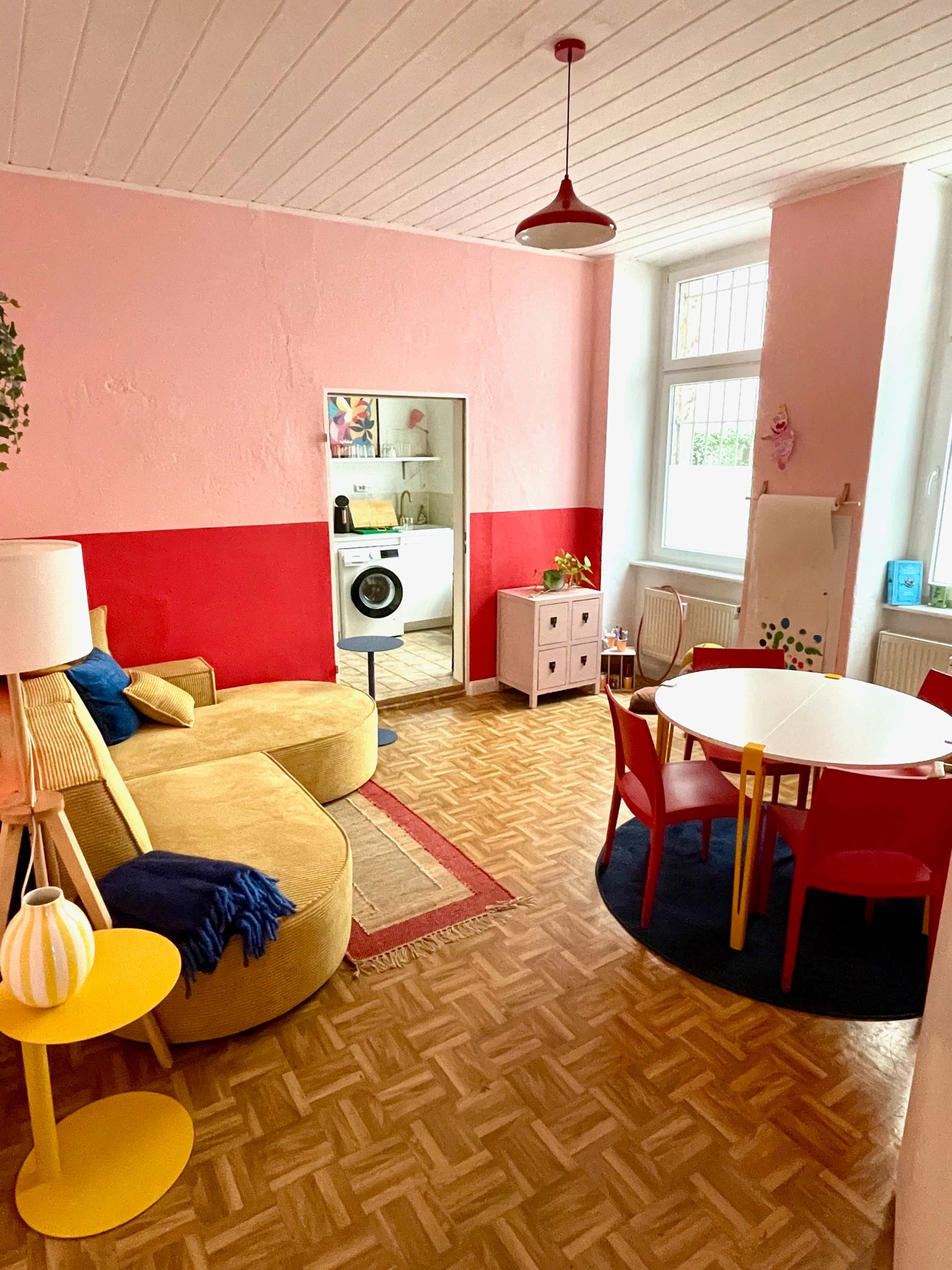 The image shows a cozy living space with a yellow sofa, a round table surrounded by colorful chairs, and a laundry area visible through a doorway.