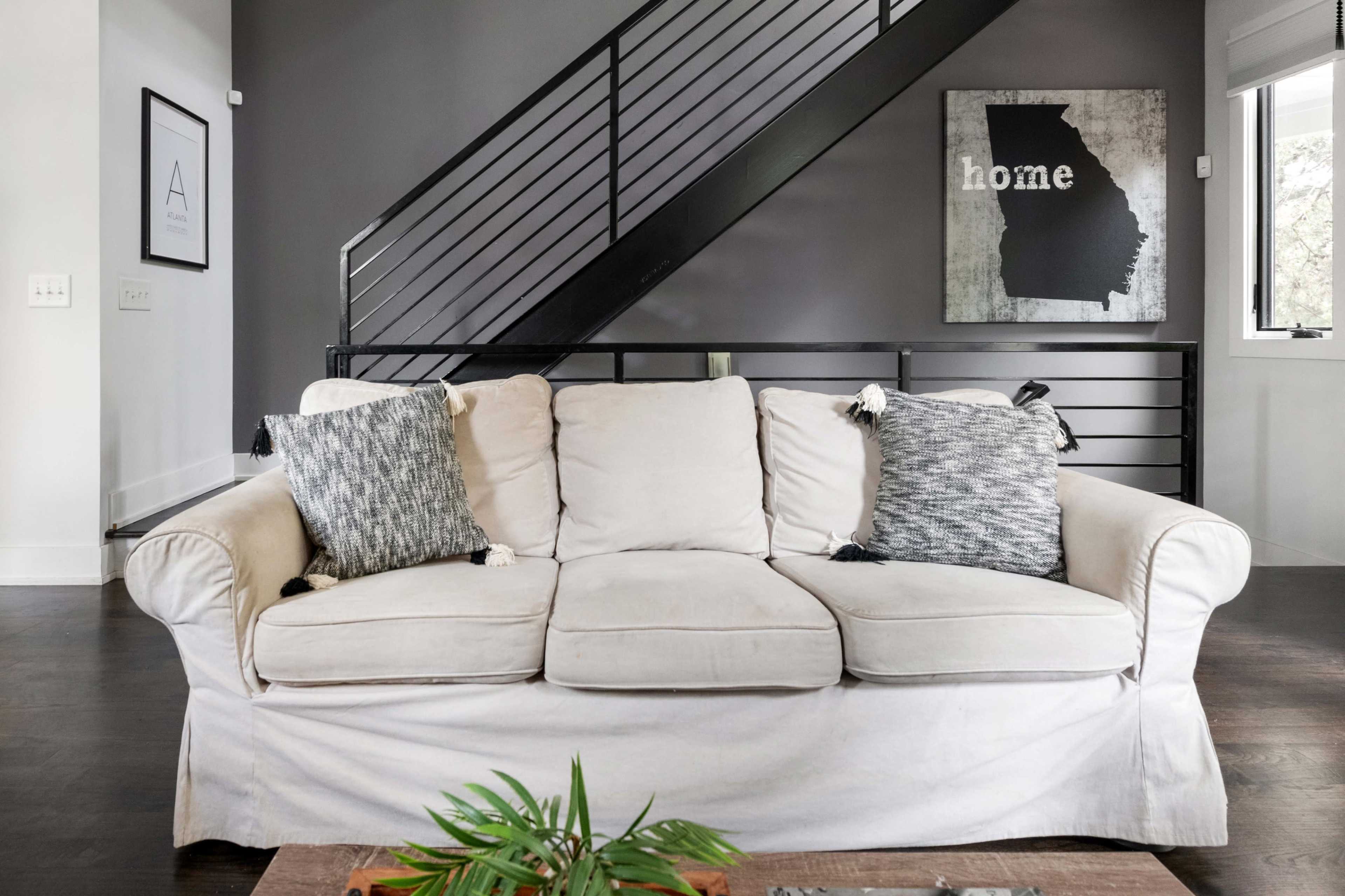 A light-colored couch with two patterned pillows sits in a living room, with a modern staircase and a wall art piece featuring the word "home" in the background.