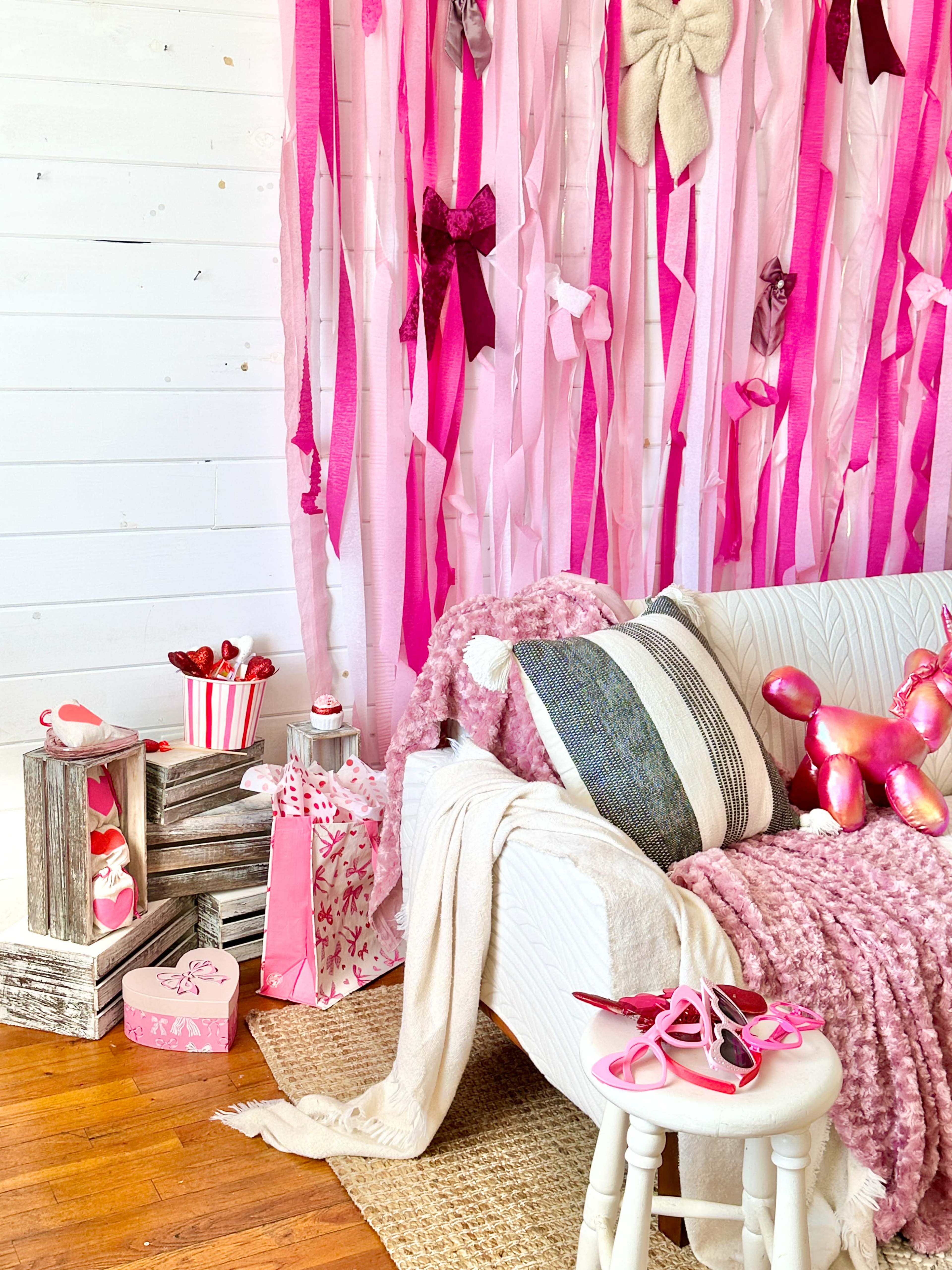 The scene features a cozy corner with a white couch adorned with pink and gray pillows, surrounded by decorations of pink ribbons, a textured throw, and various pink-themed items, including gift boxes and a small popcorn bucket.