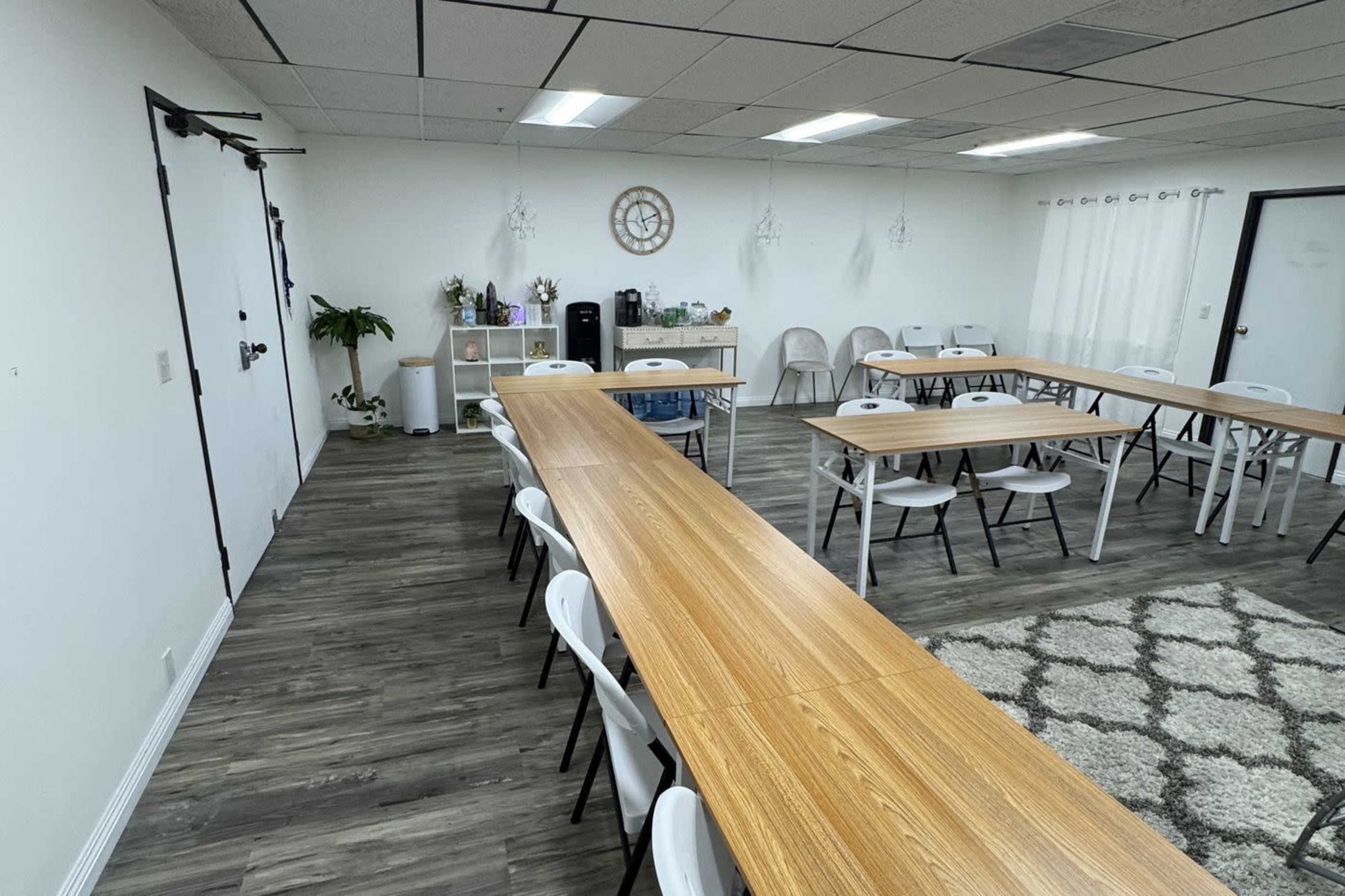 The image shows a modern meeting room with a long wooden table set up for seating, white chairs arranged neatly around it, and a wall adorned with a clock and minimal decor.