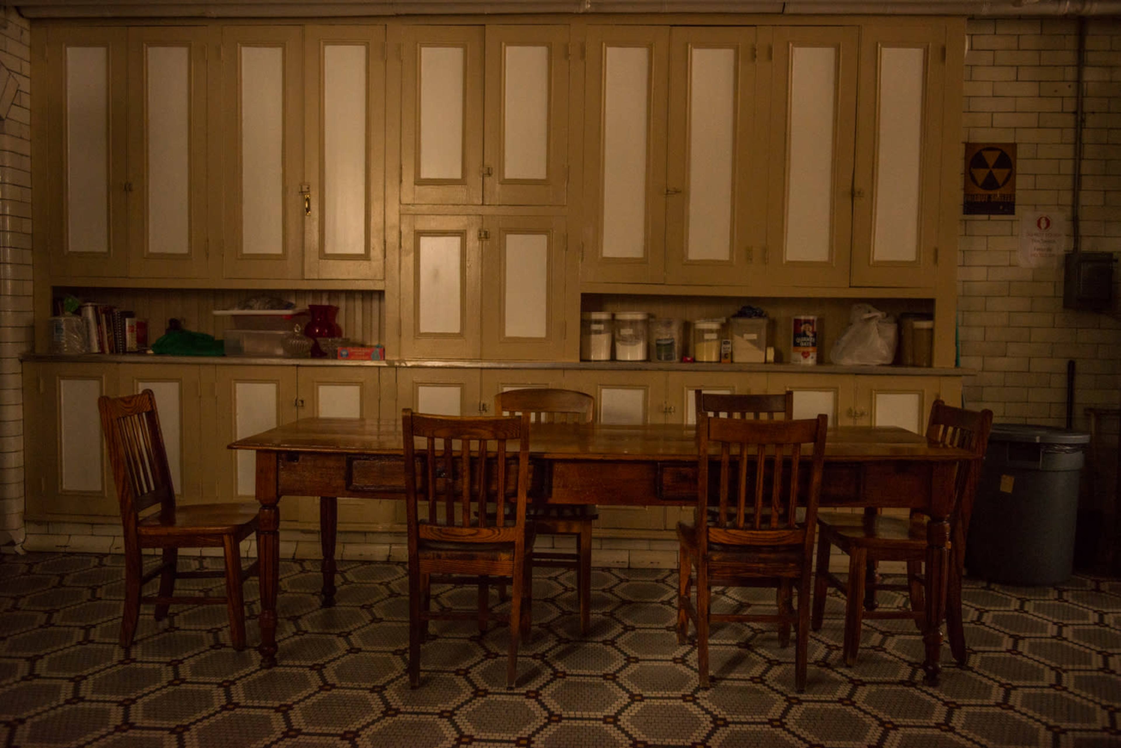 A long wooden table surrounded by four wooden chairs sits in a dimly lit room with a cabinetry wall in the background.