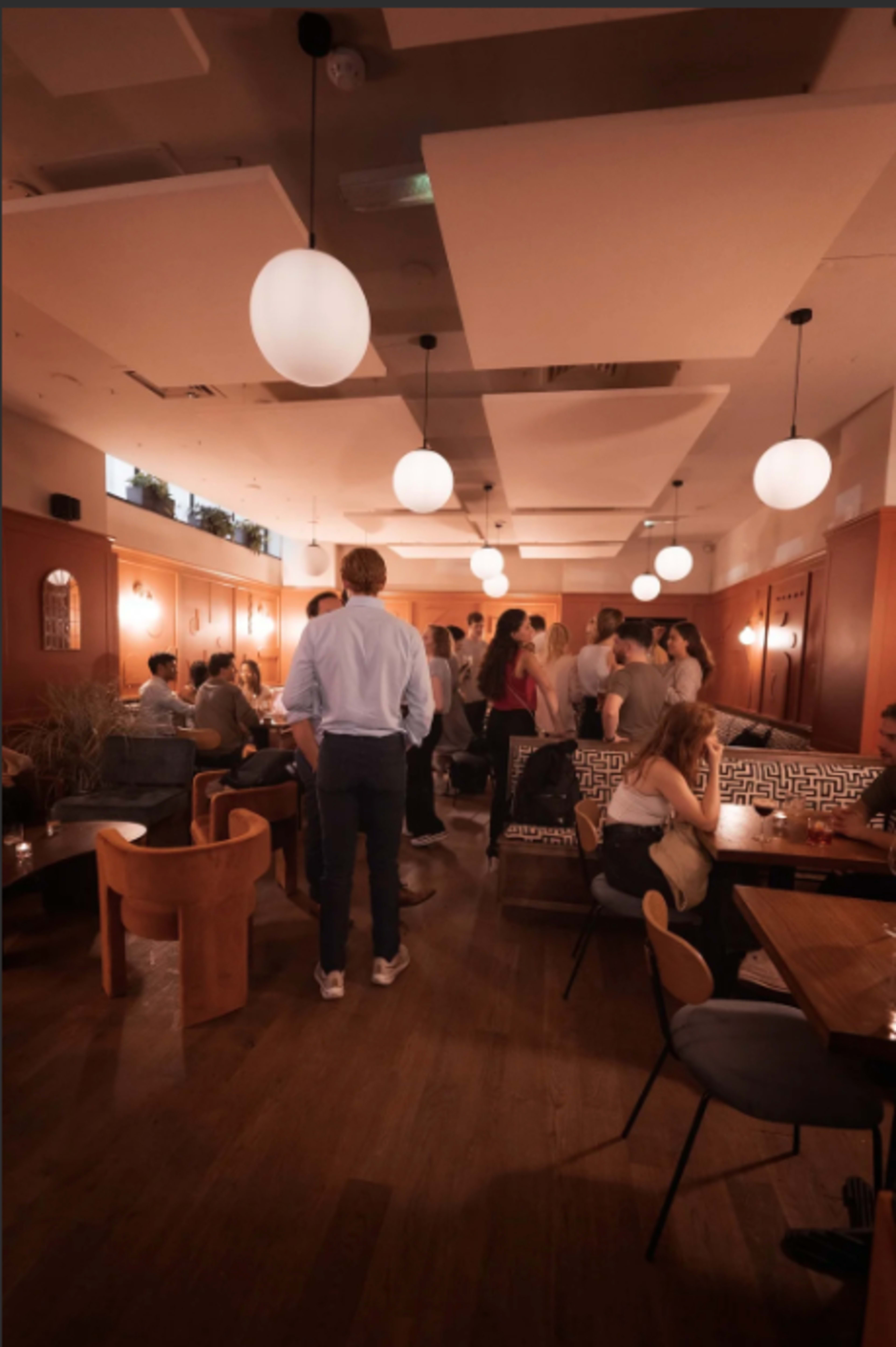 The image shows a bustling indoor restaurant with multiple groups of people socializing, warm lighting, and a mix of seating arrangements.