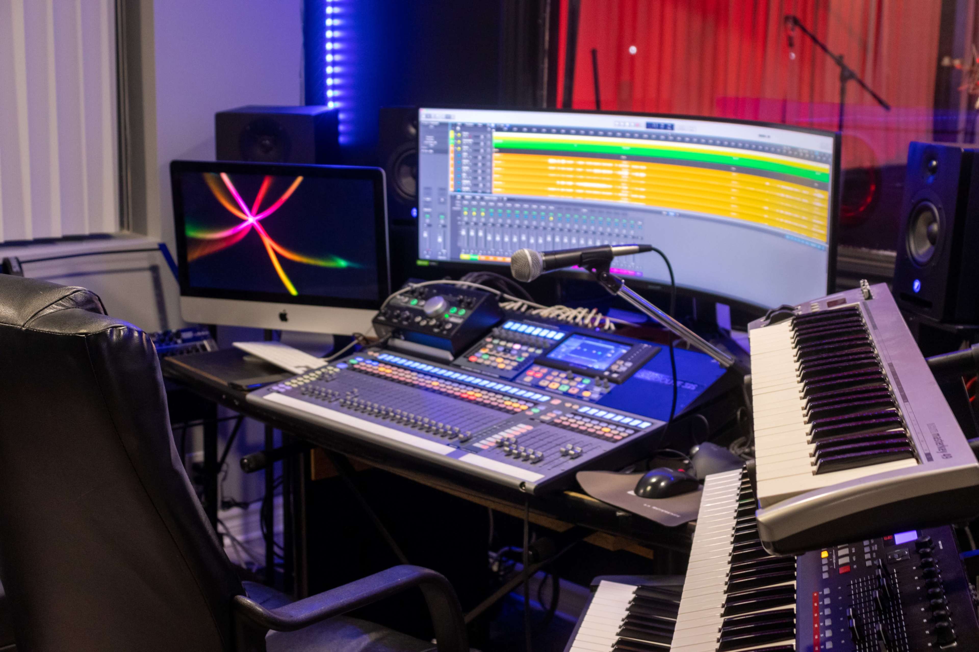 A recording studio setup features a large computer monitor displaying audio tracks, a microphone on a mixing console, and multiple musical keyboards.