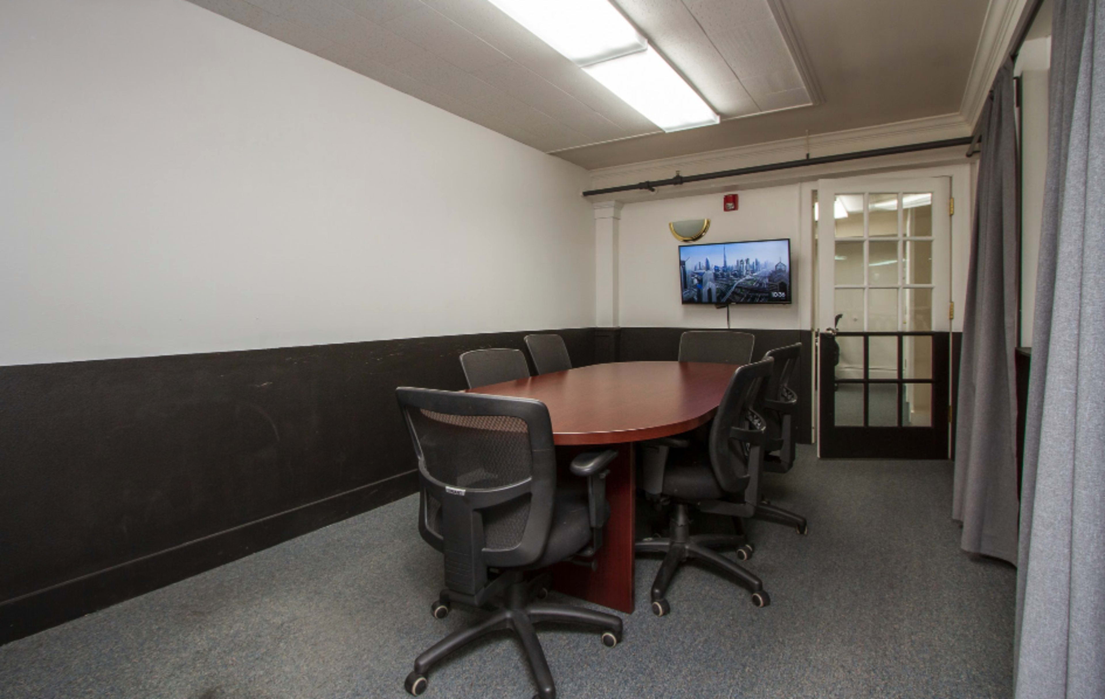 A conference room features a large wooden table surrounded by black office chairs, with a television mounted on the wall and a set of double doors leading to another area.