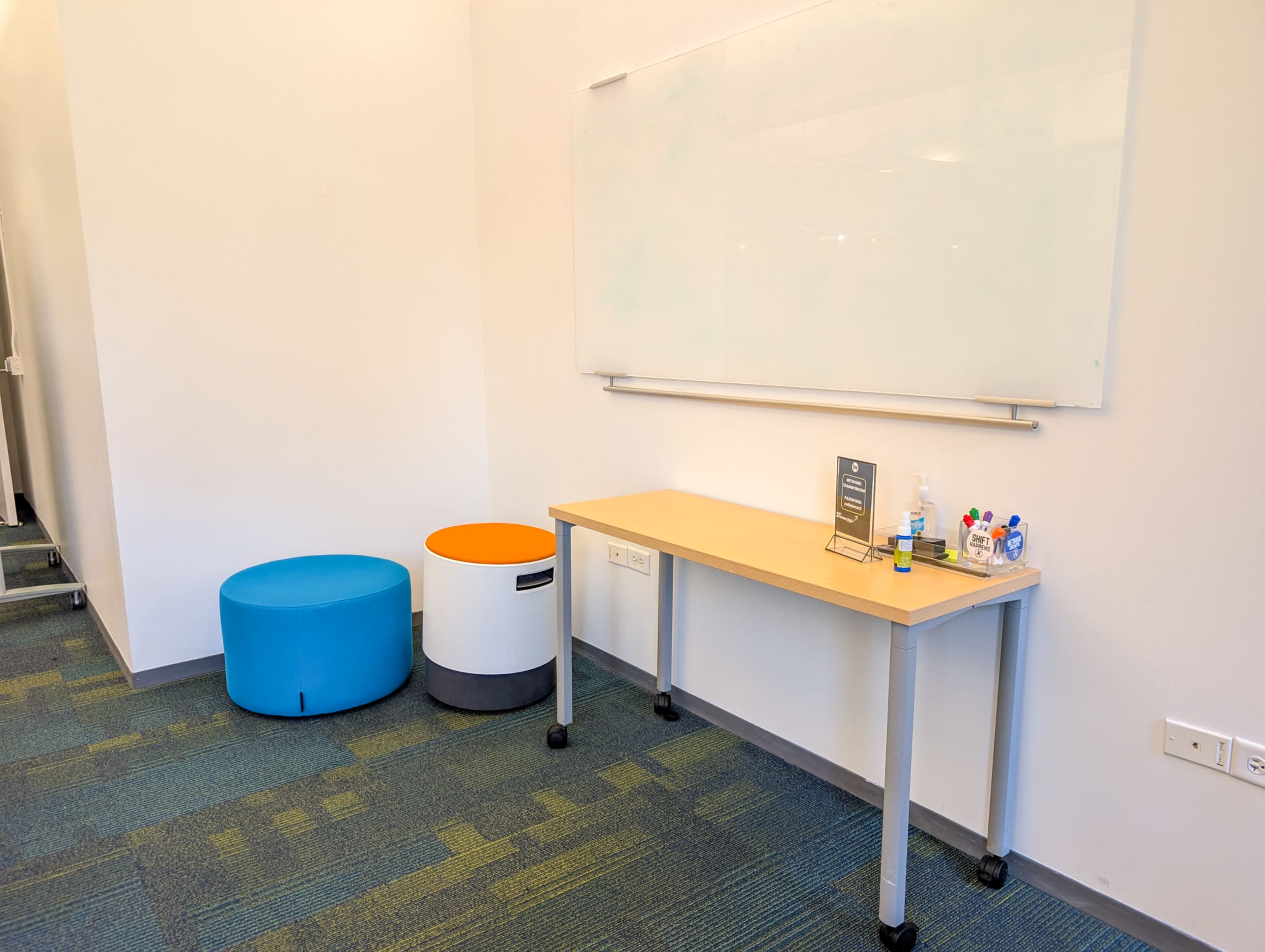 The image shows a minimalist office space featuring a small table on wheels and two cylindrical seating options, one blue and one orange, beside a whiteboard on the wall.