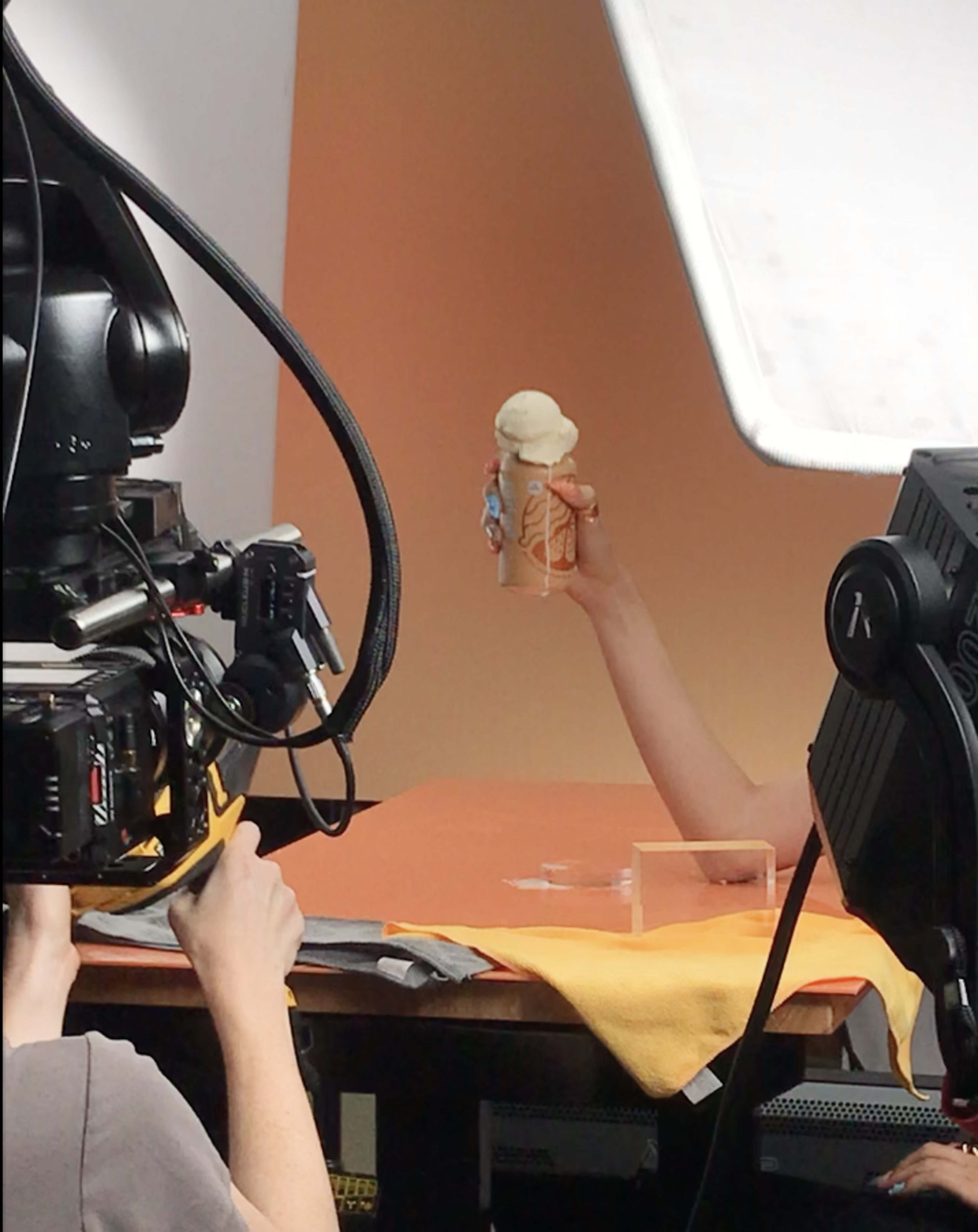 A hand holds an ice cream cone in front of a camera setup with bright lighting during a photoshoot.